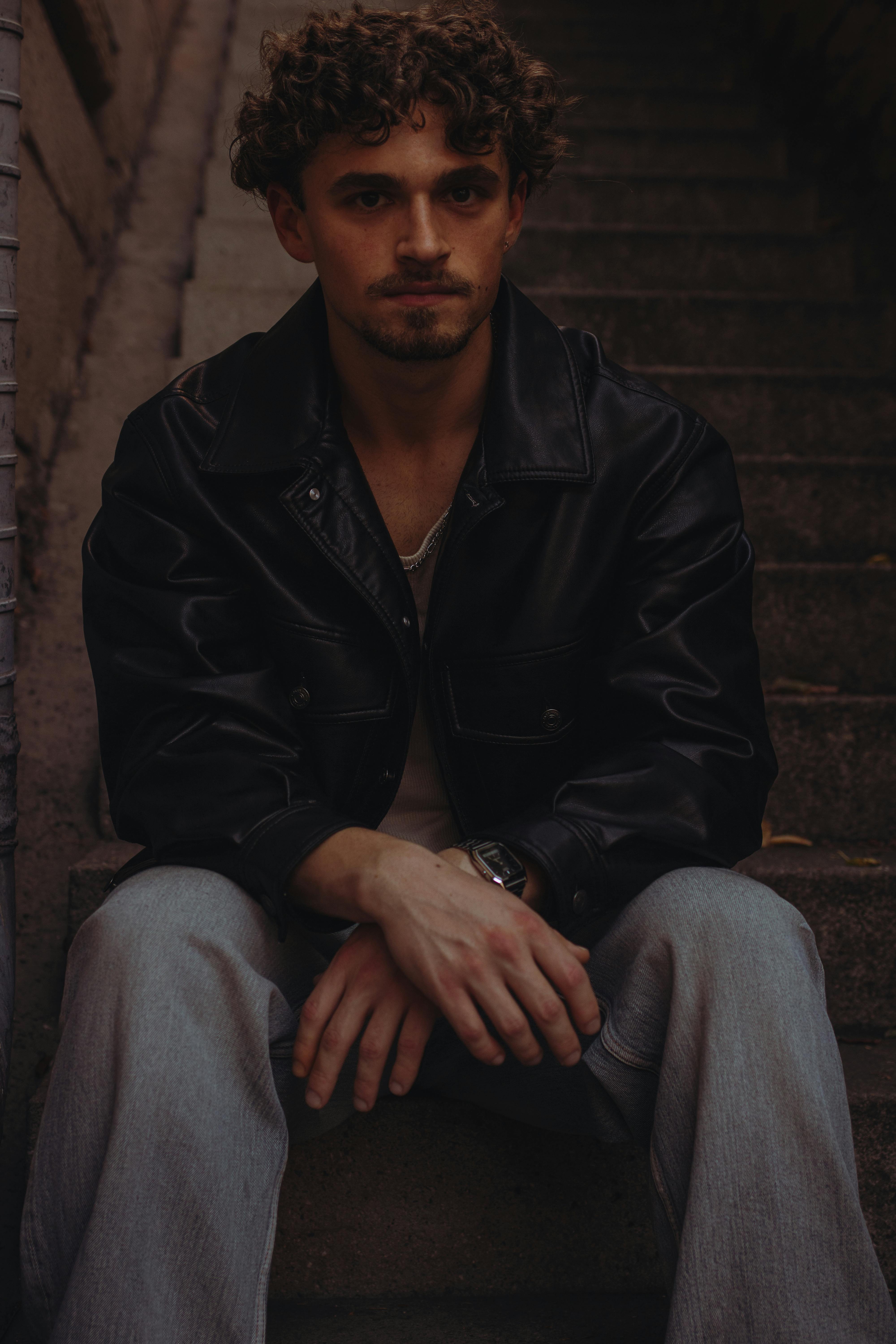 A man with curly hair sits on stairs wearing a leather jacket, portraying a thoughtful urban vibe.