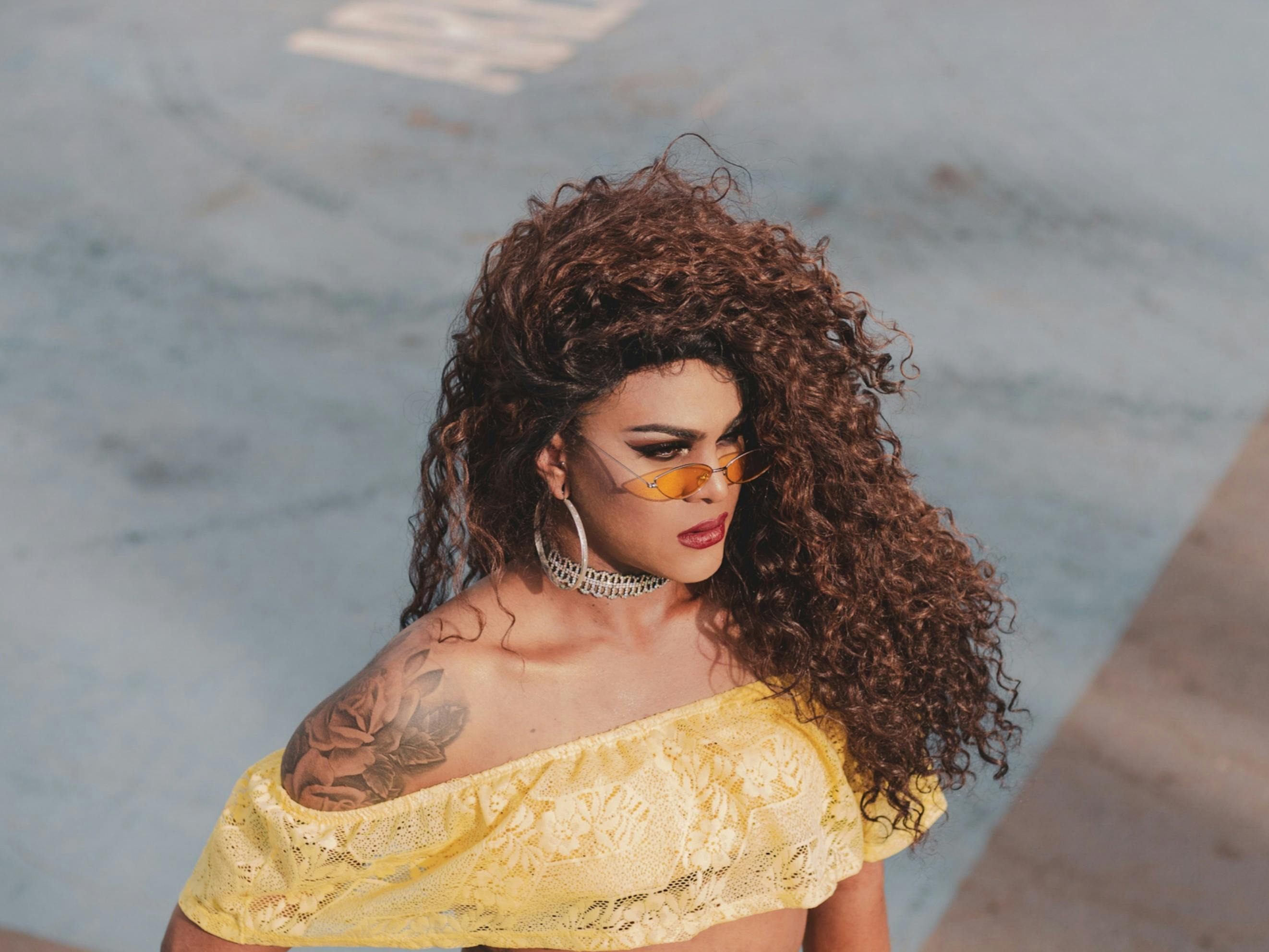 一名卷发女子身穿黄色蕾丝上衣，戴着太阳镜，身上有纹身，正在户外摆姿势