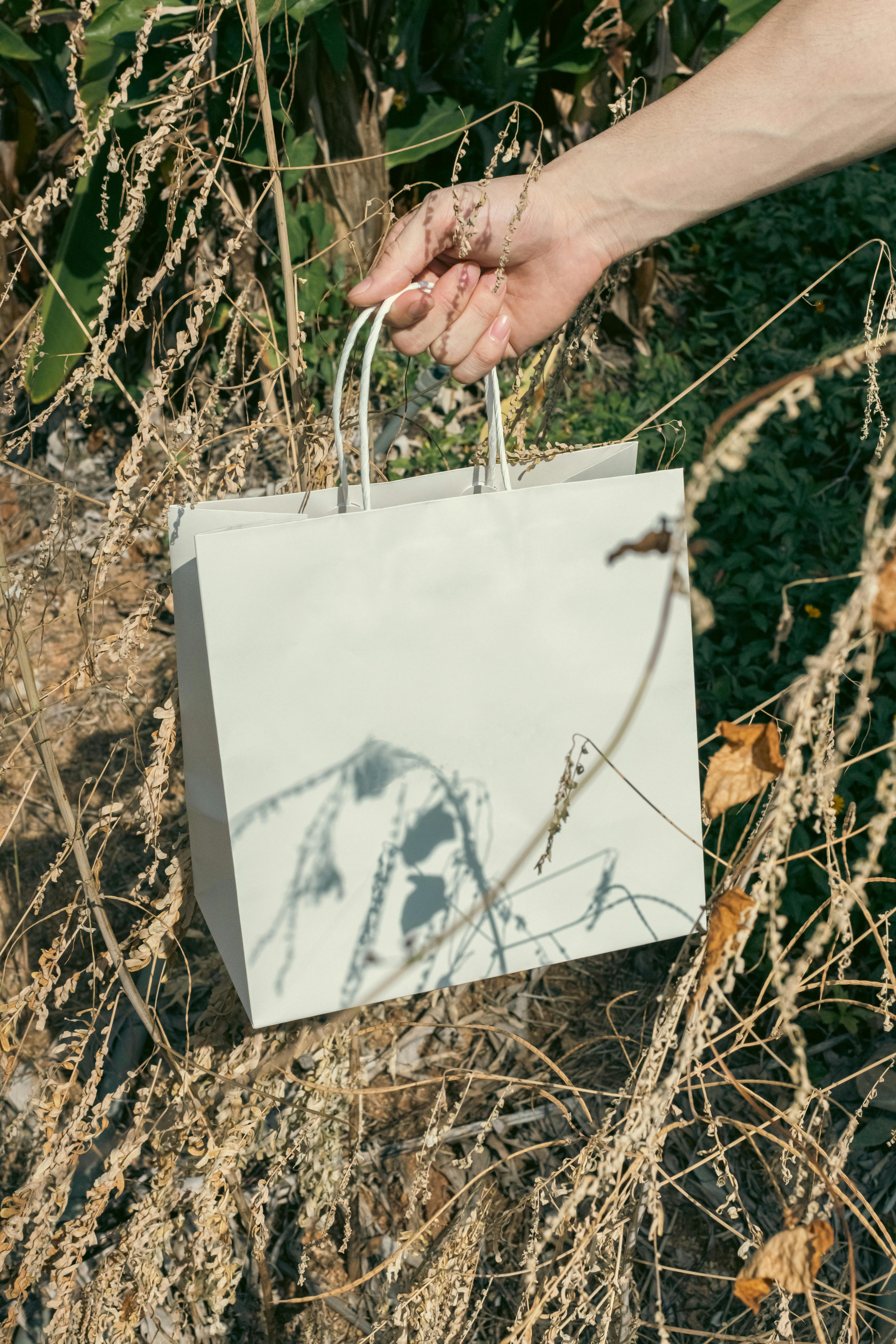 A hand holding a reusable paper bag among dry grass, promoting sustainability.