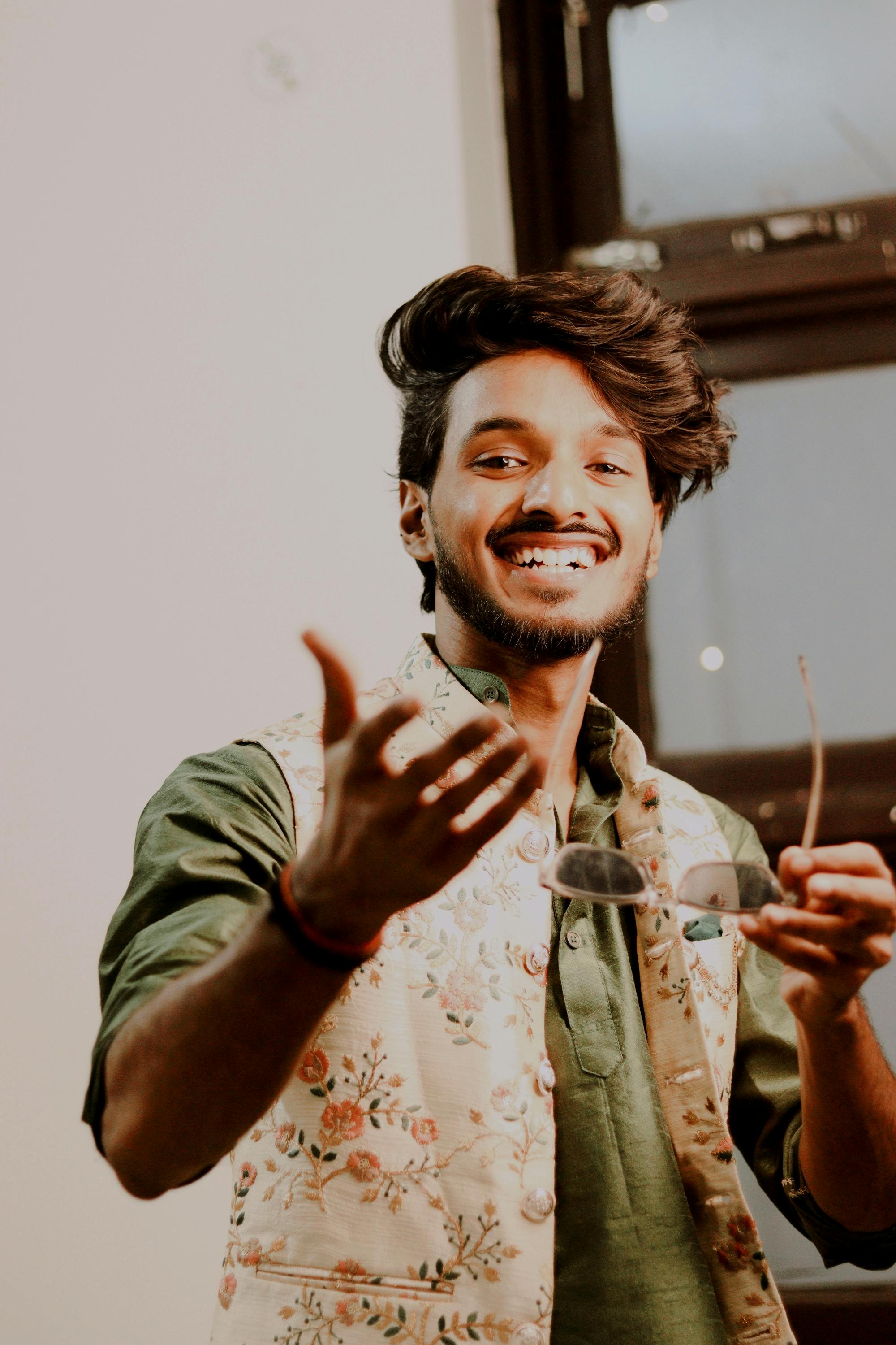 Smiling man with a beard and mustache in a floral vest holding glasses. Indoors setting in New Delhi, India.