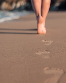 Close-up of a woman's legs walking barefoot on a sandy beach, leaving footprints behind.