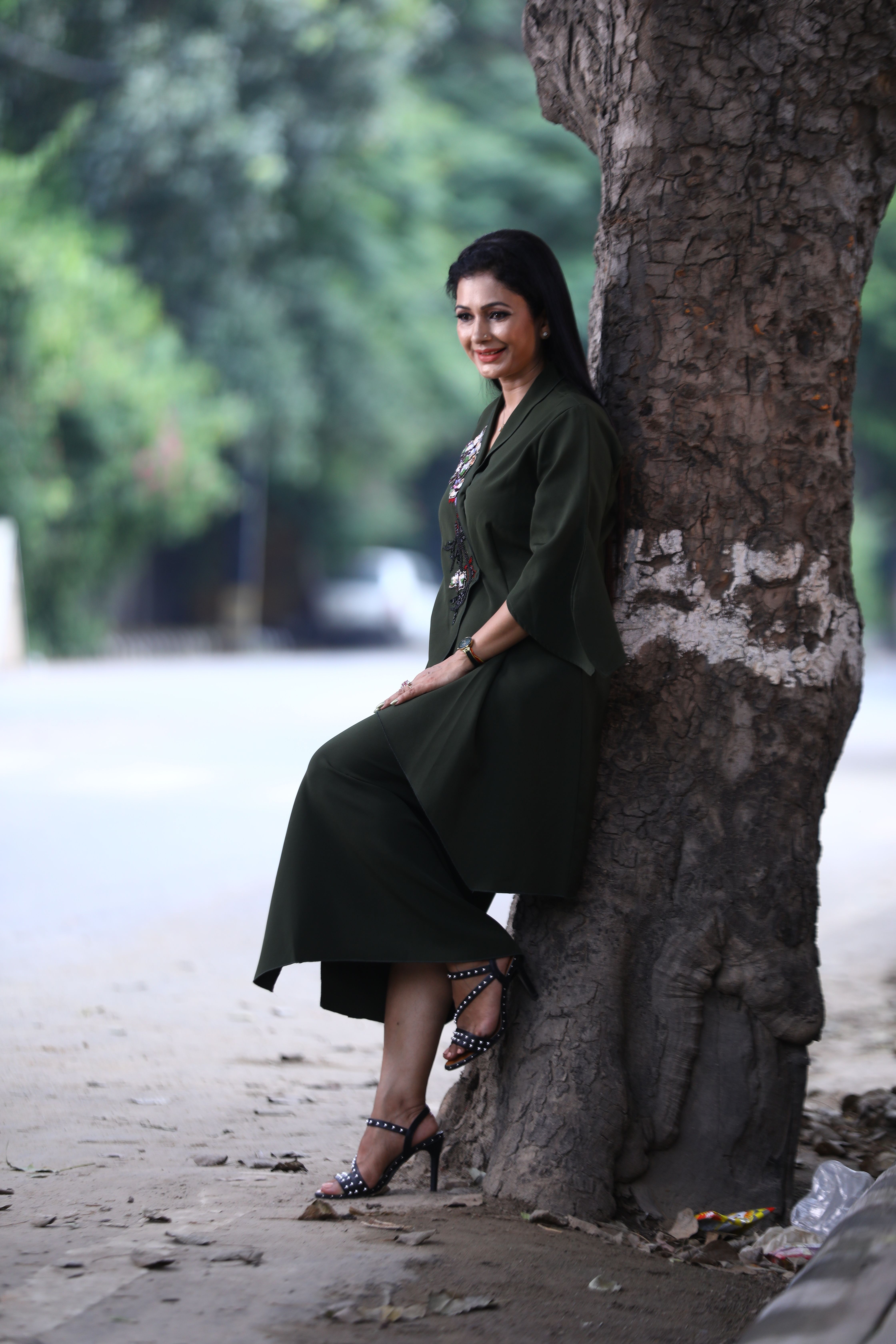 A fashionable woman in a green dress smiles while leaning against a tree outdoors, exuding elegance.