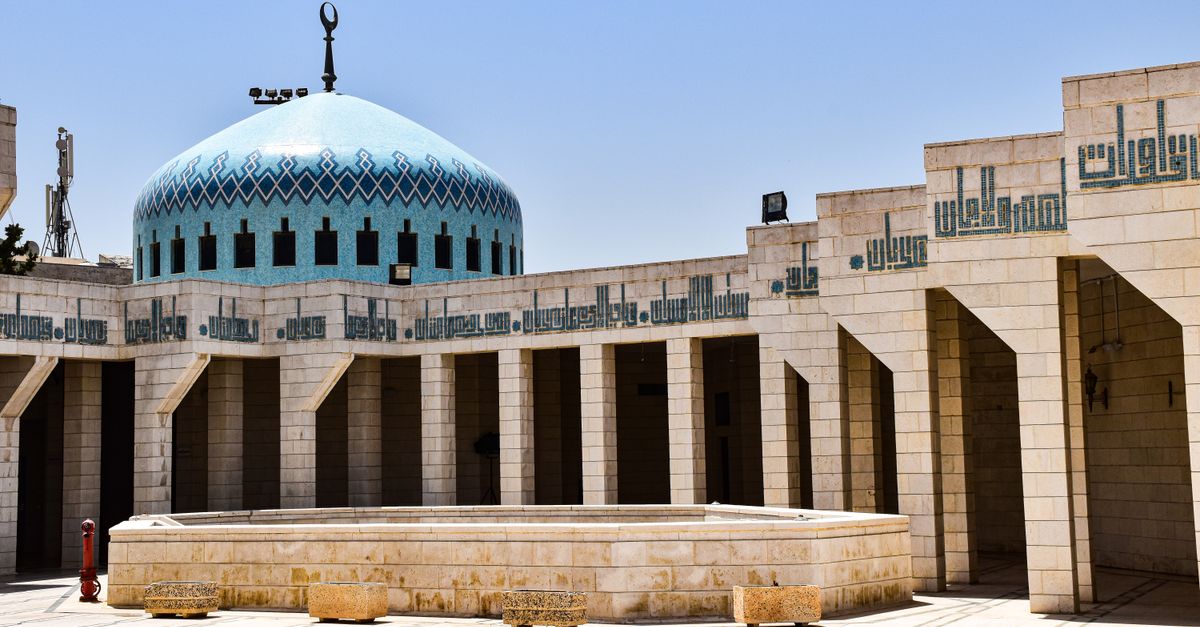 The official Jordanian flag ripples in the wind against a clear blue sky outside a government building.
