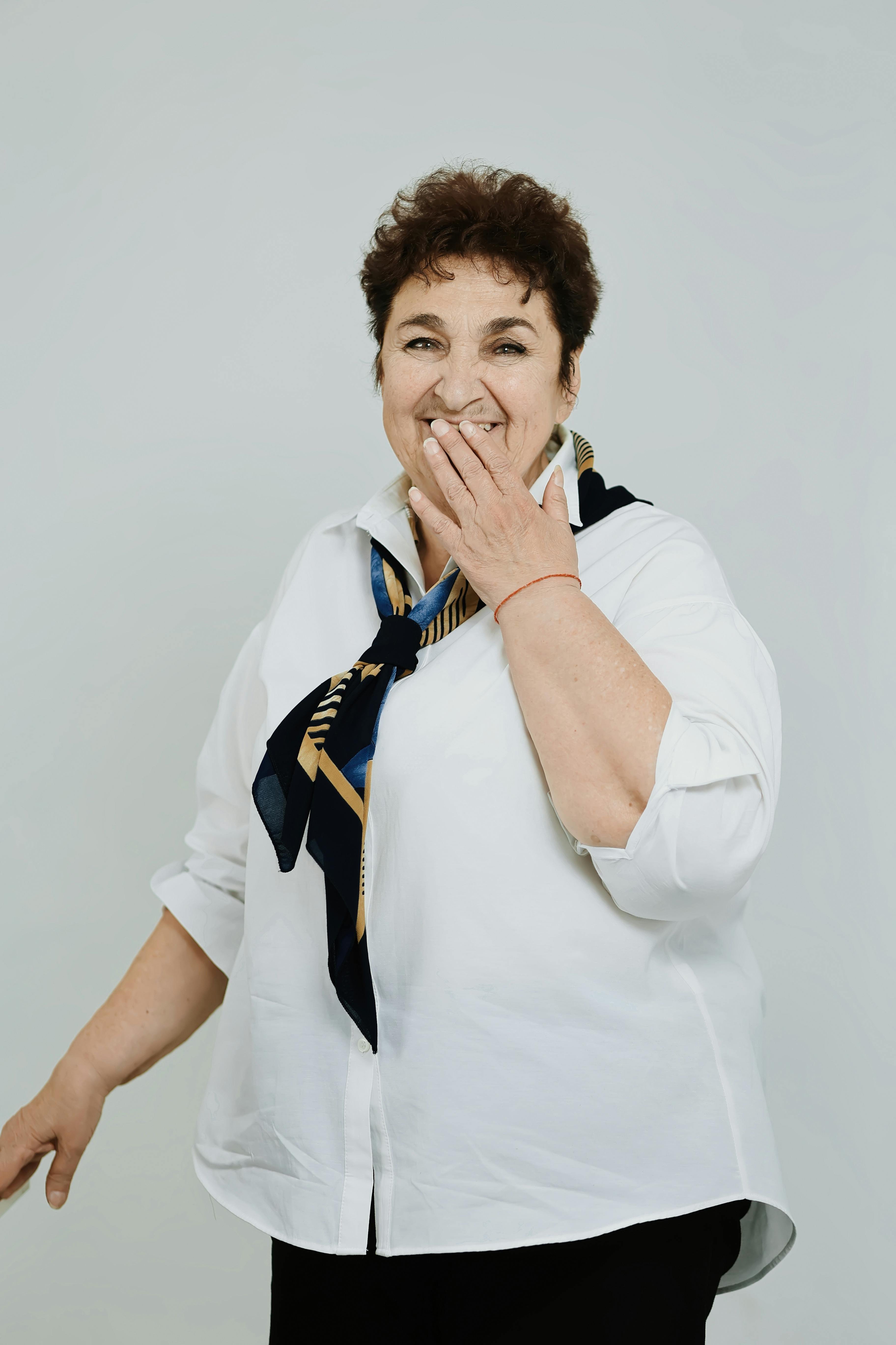 Stylish senior woman in white shirt and scarf laughing joyfully against a plain background.