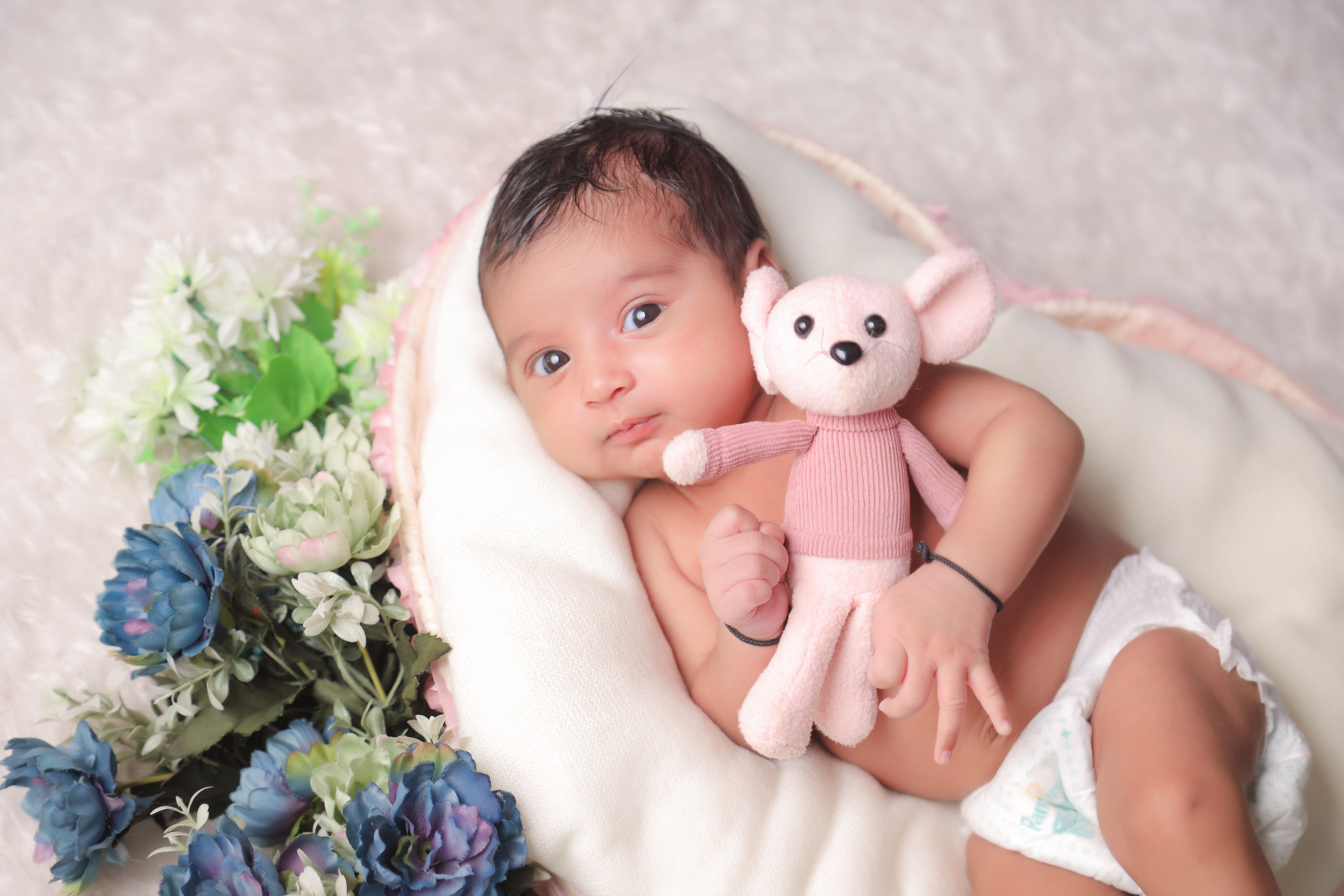 Cute baby holding a teddy bear, lying on a cozy blanket surrounded by flowers.