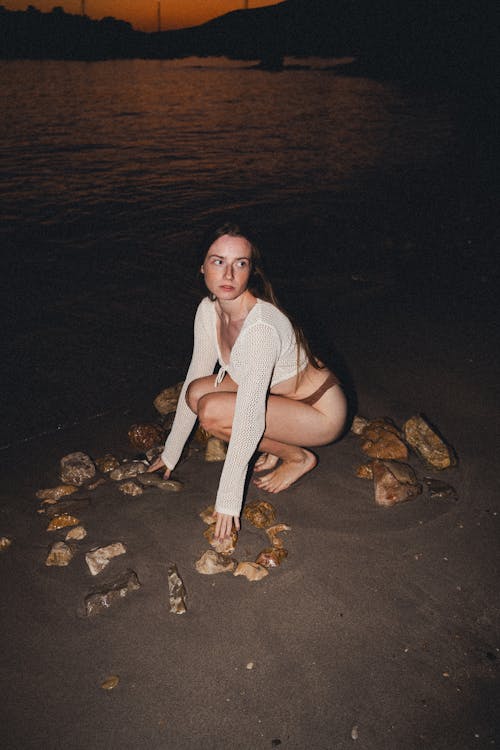 Brunette Woman in White Top Crouching on Lakeside in Evening