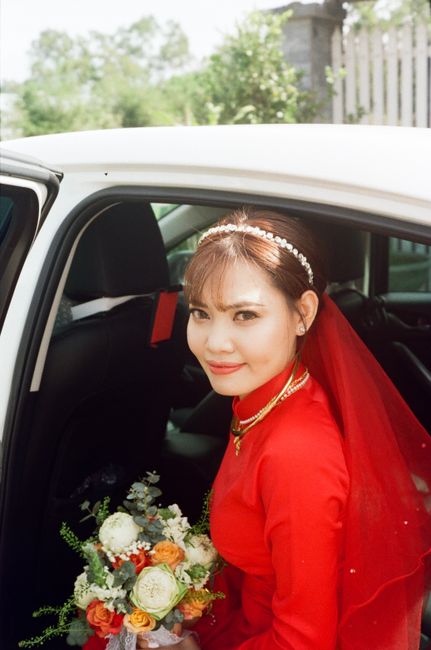 Asian bride in red dress sitting in car holding a bouquet, smiling.