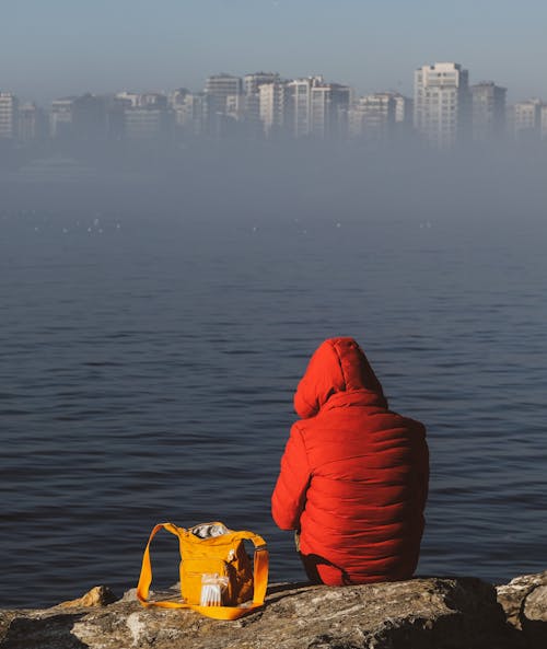 Man Wearing a Red Jacket with a Hood, Sitting on a Riverbank, and City Buildings in Mist