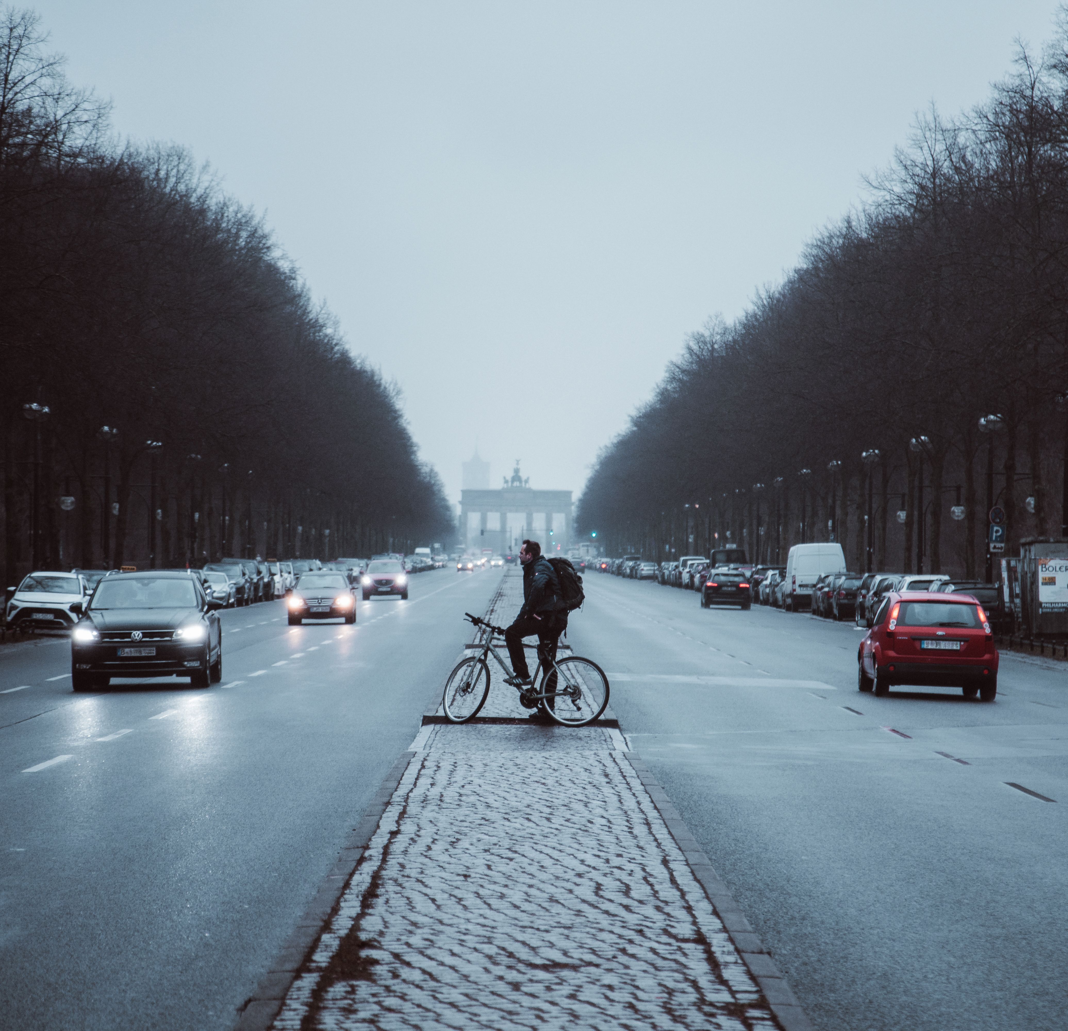 Cyclist rides through foggy Berlin street with Brandenburg Gate in view, amidst light traffic.