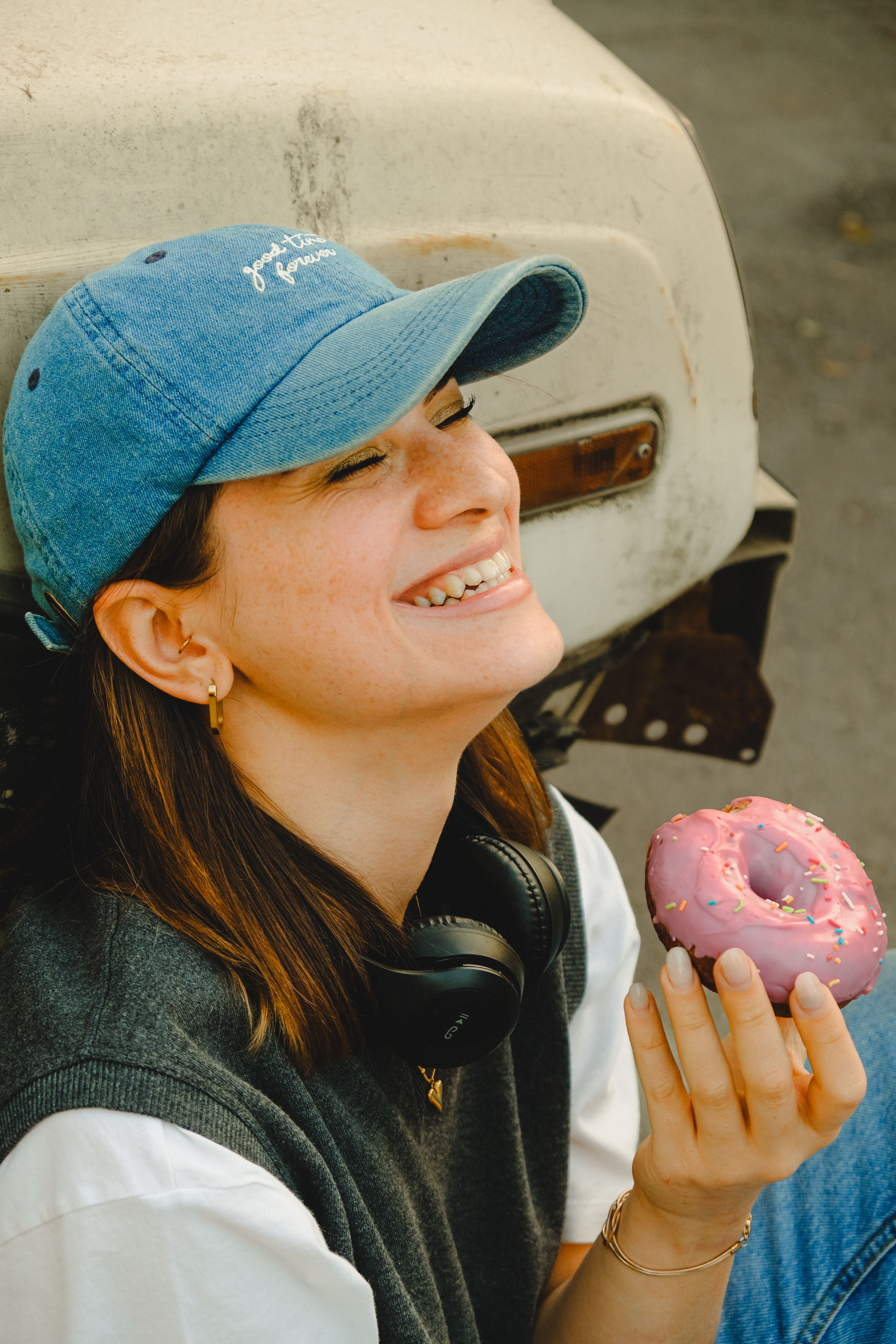 A joyful woman wearing a denim cap smiles while savoring a pink glazed donut outdoors.