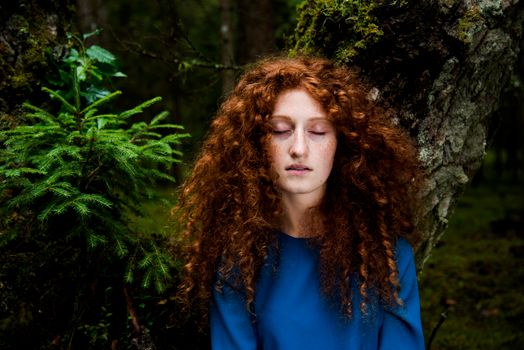Portrait of a redhead woman with eyes closed amidst vibrant green foliage in a forest.