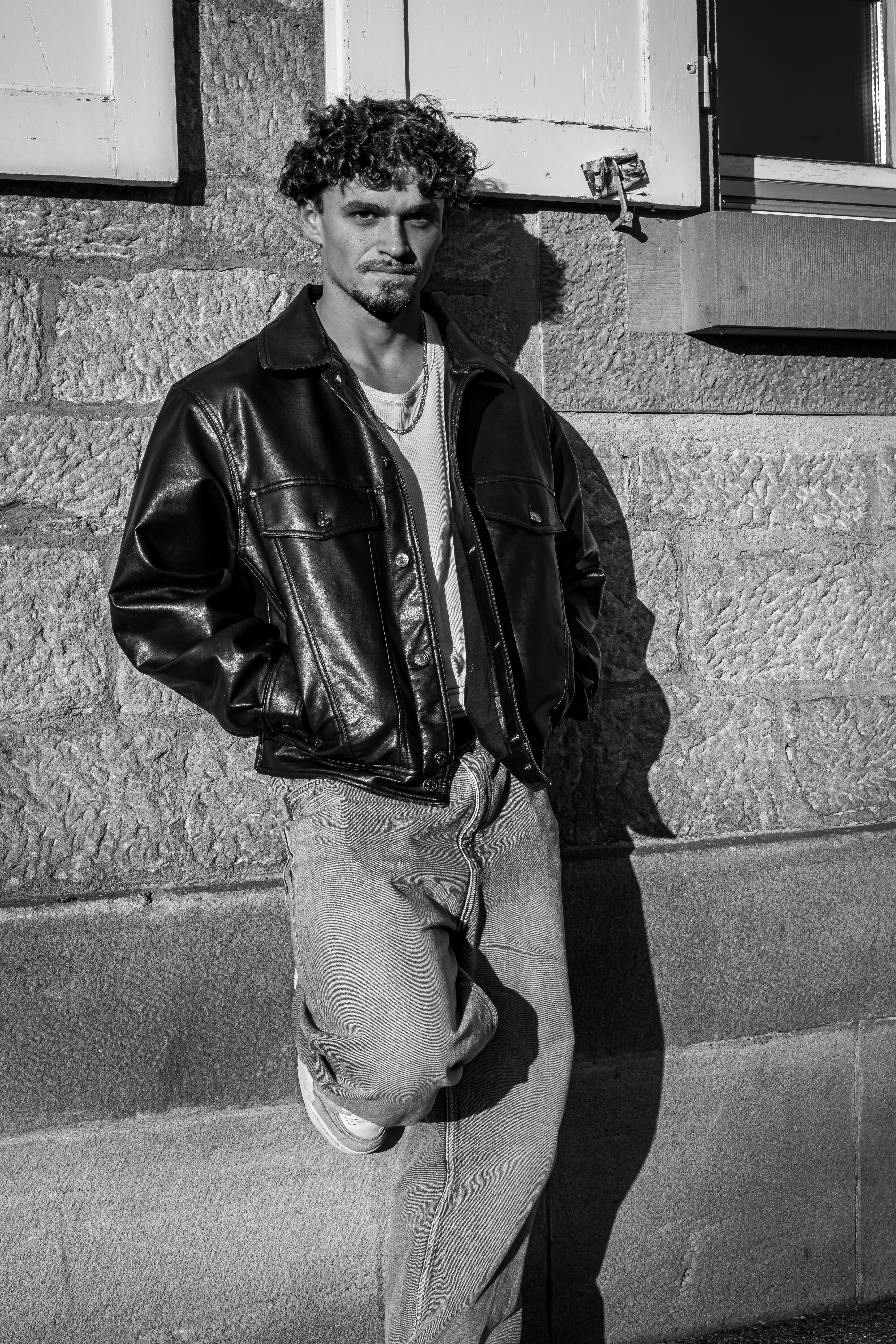 Black and white portrait of a man in a leather jacket leaning against an urban wall.
