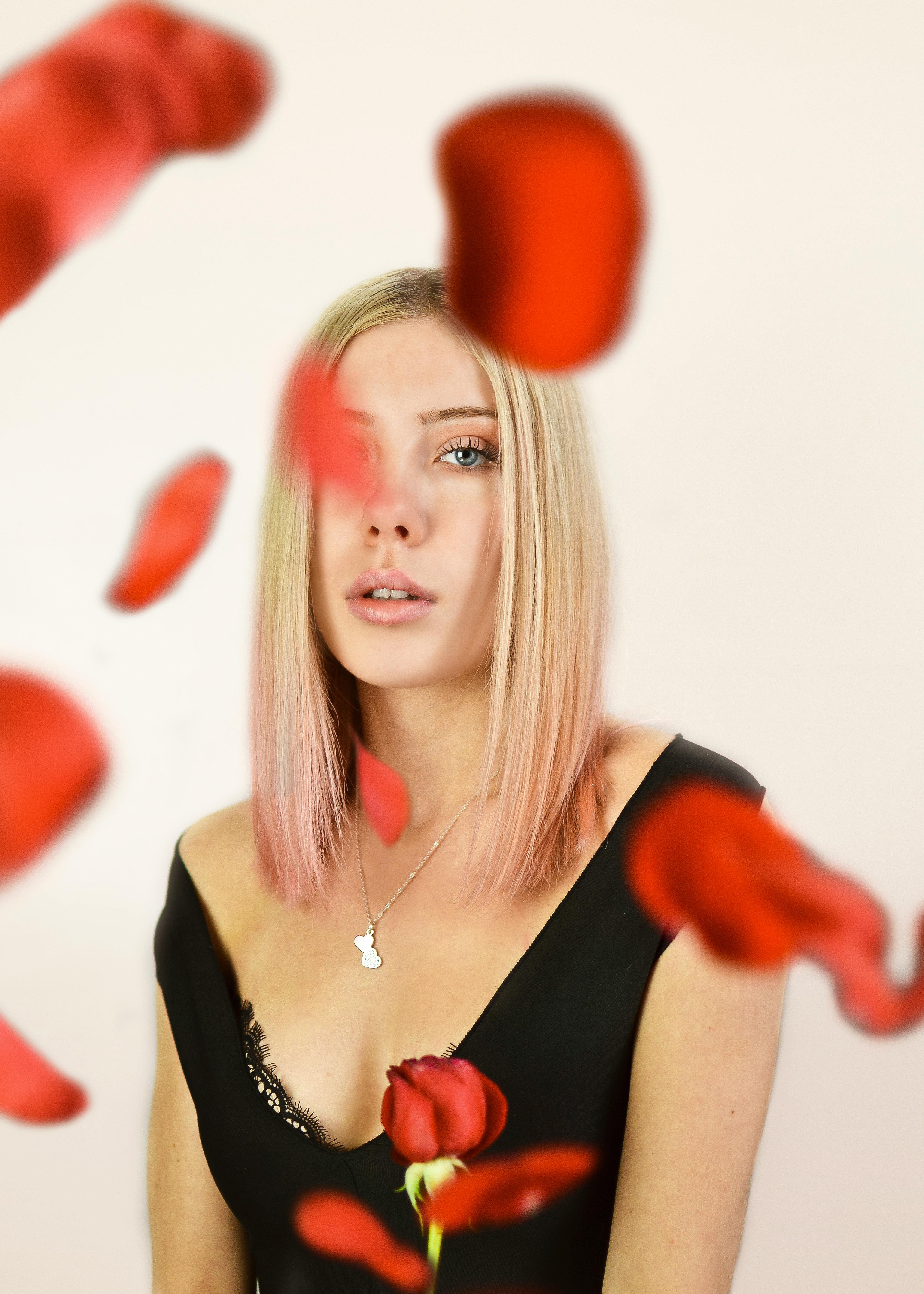 一位金发女子身着黑色连衣裙，周身环绕着鲜艳的红色玫瑰花瓣