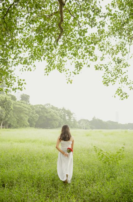A woman in a white dress holding red flowers walks across a vibrant green field under a tree.