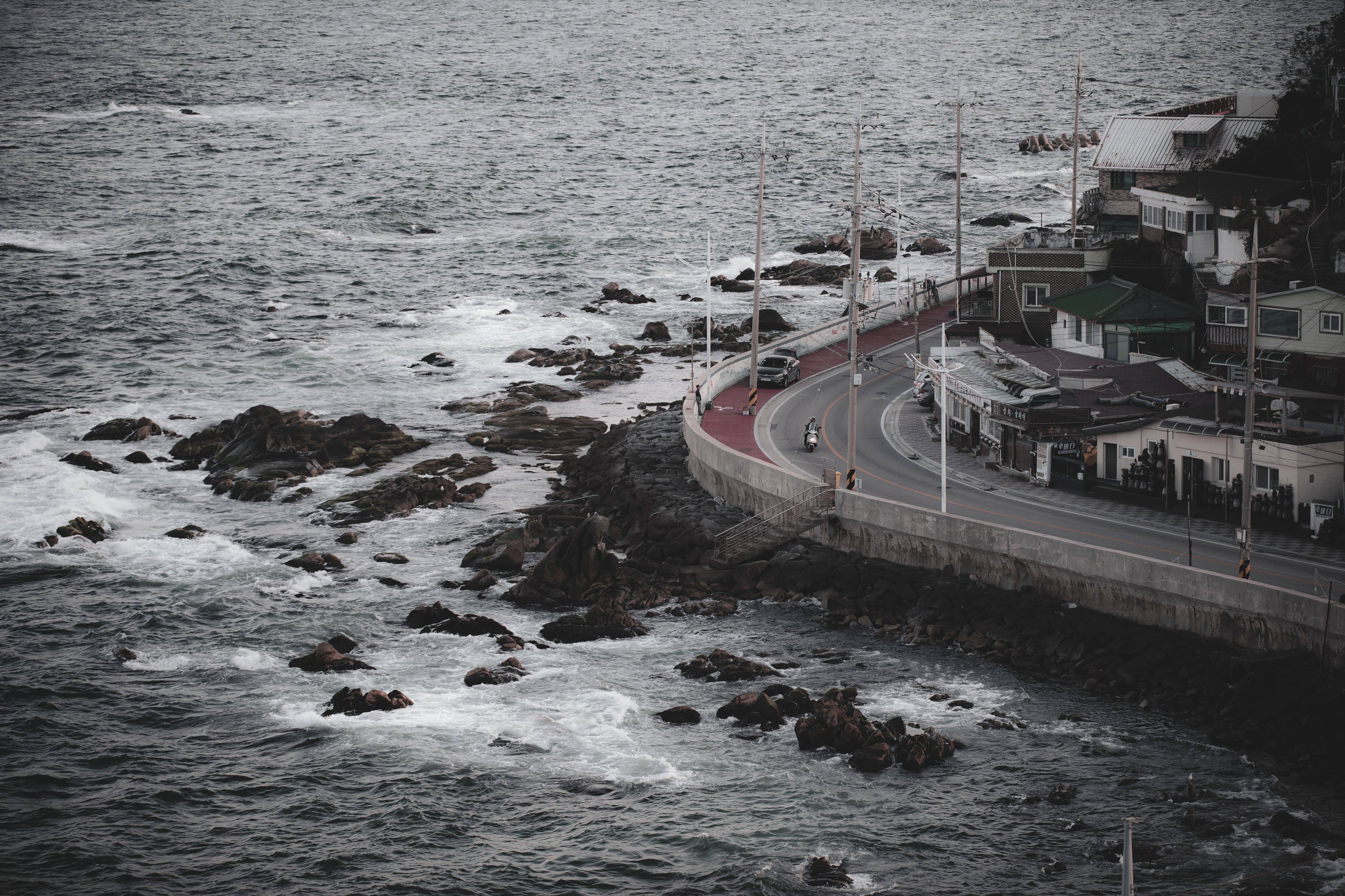 Aerial view of a coastal road and town in Gangneung, South Korea, with rocky shore and sea.
