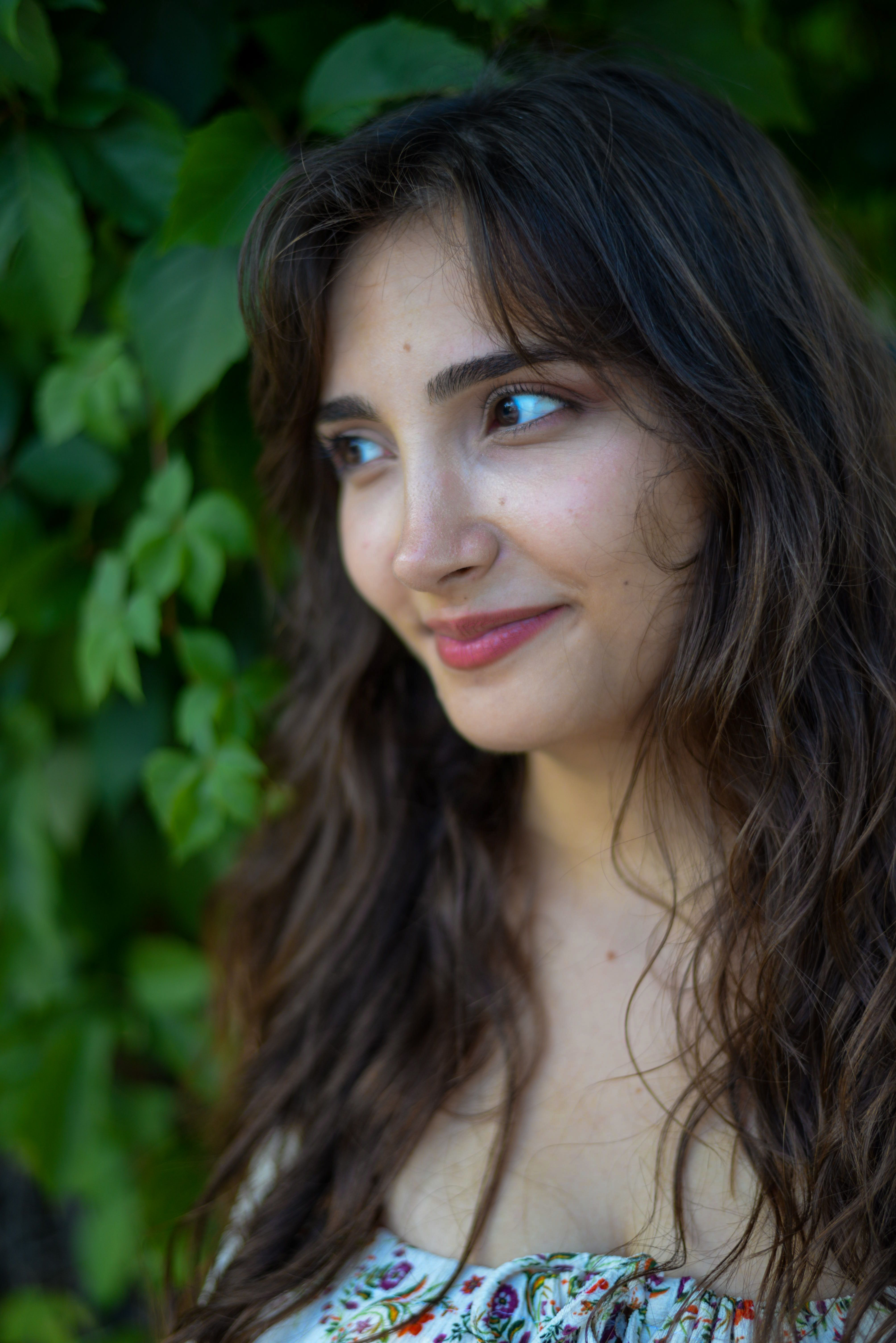 A young woman with brunette hair smiles outdoors among green leaves.