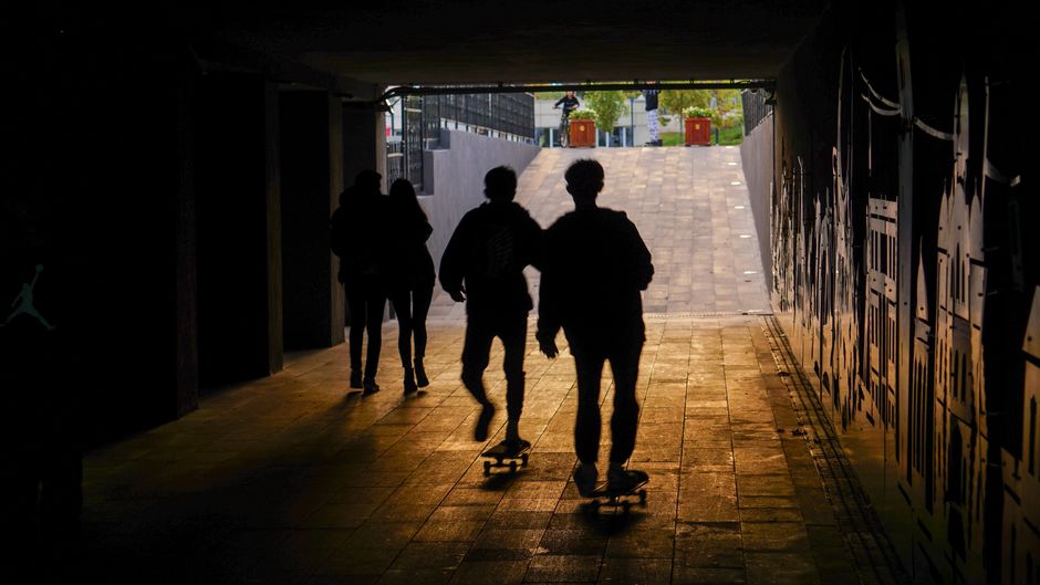 Siluetas de skaters emergiendo de un túnel tenuemente iluminado hacia la luz del día de las calles urbanas.
