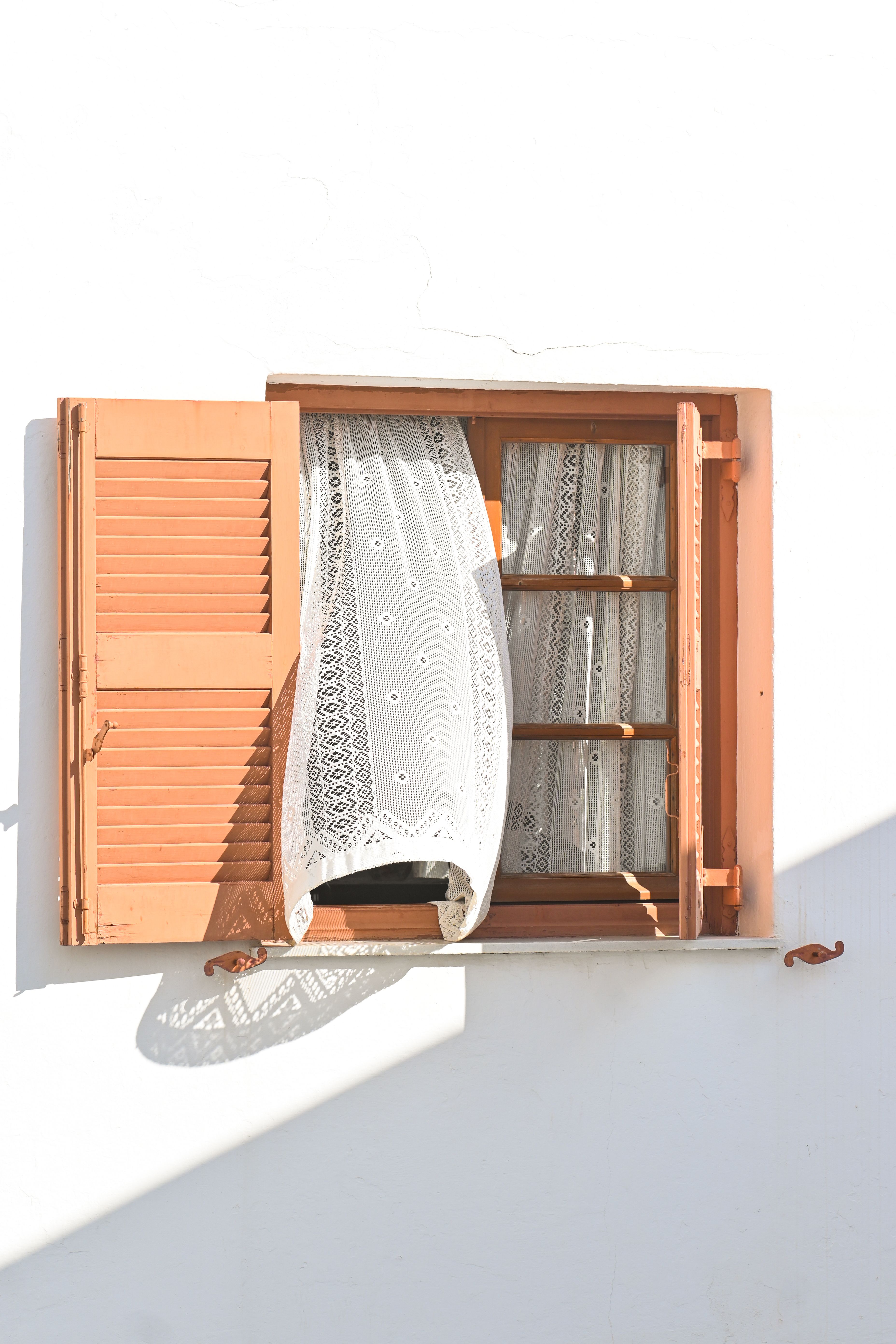 Sunlit wooden window with lace curtain blowing gently, featuring pastel shutters on a white wall.