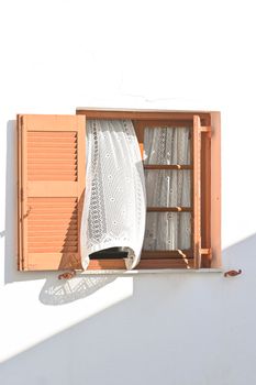 Sunlit wooden window with lace curtain blowing gently, featuring pastel shutters on a white wall.