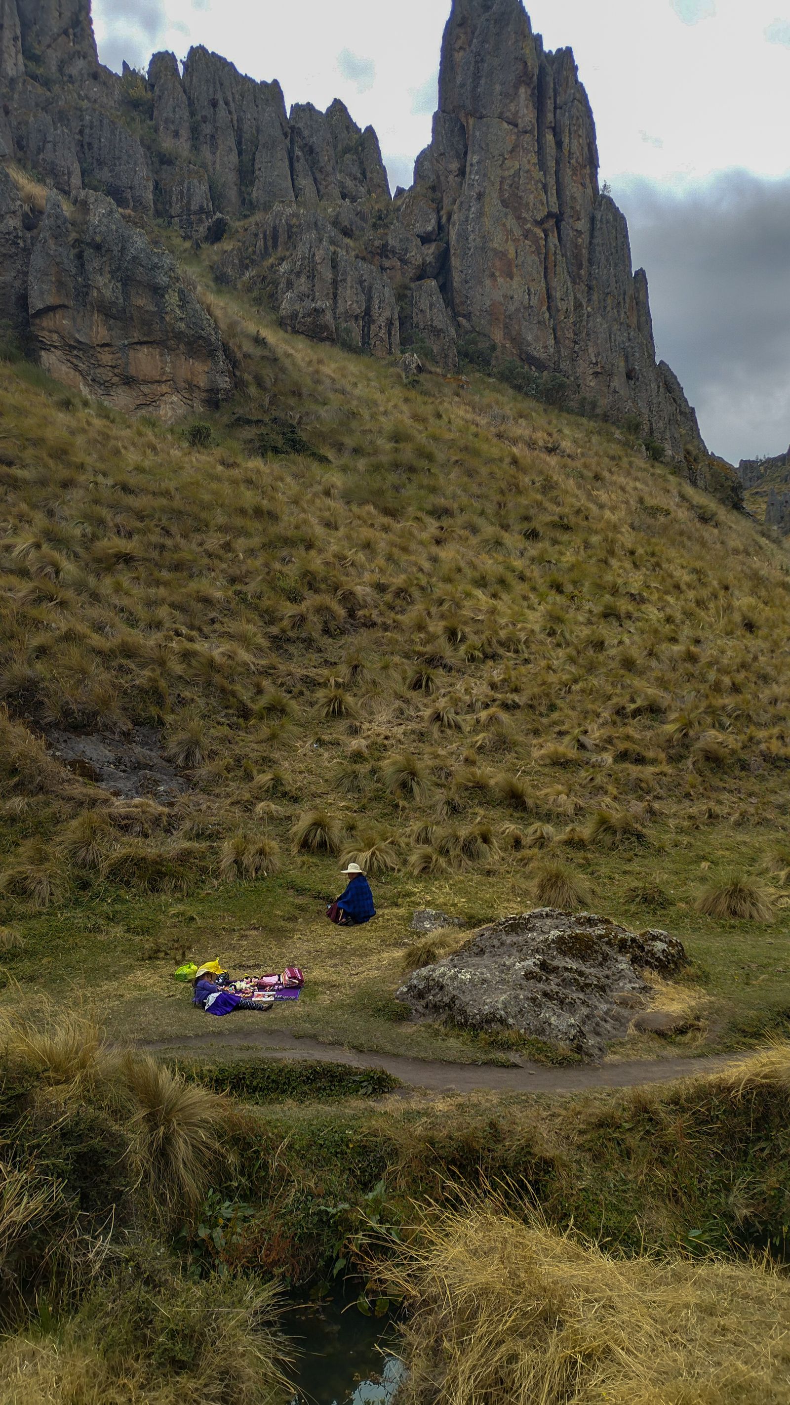 Ladera de pasto seco al pie de altas paredes de roca gris, con una persona sentada en el prado junto a un sendero