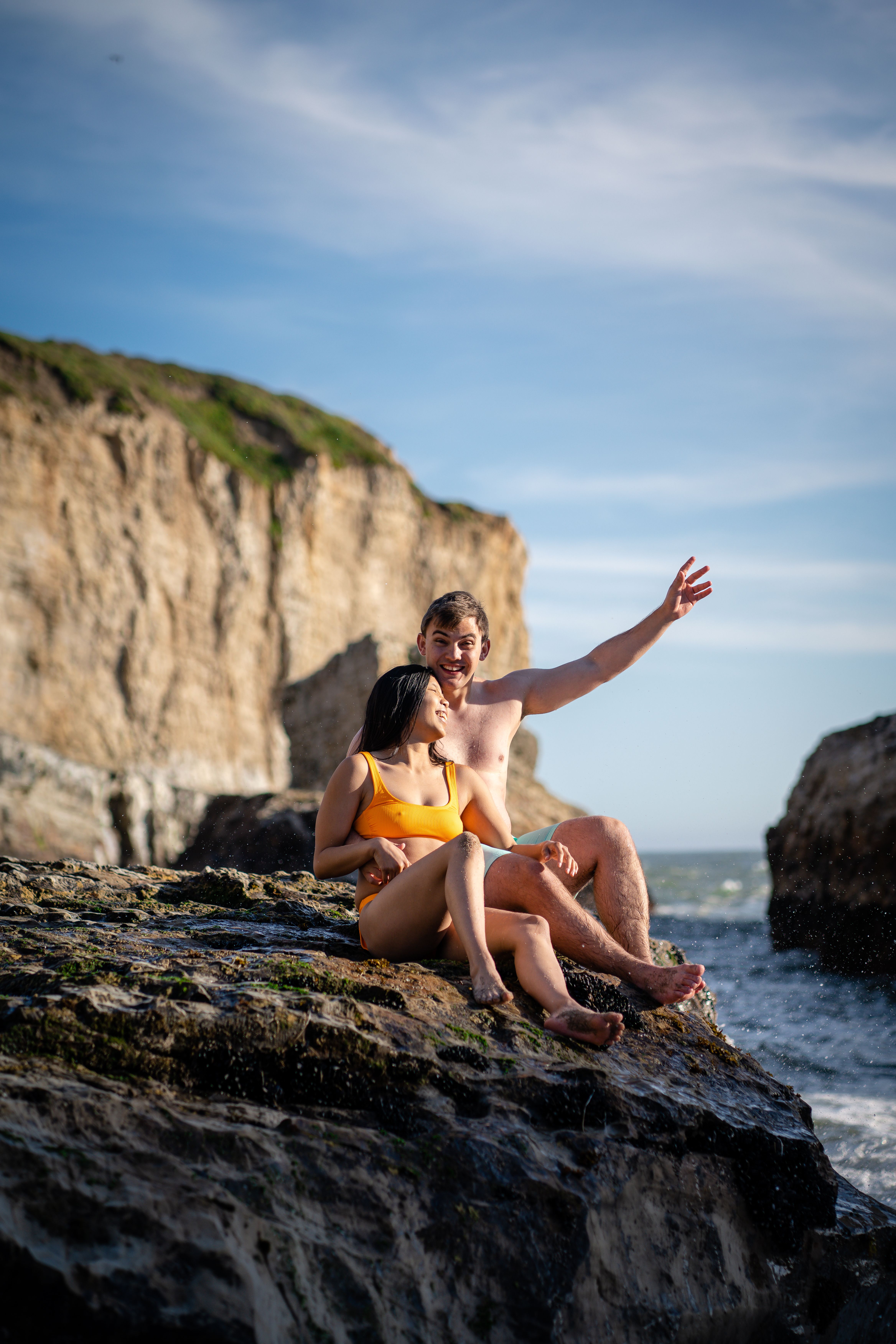 A happy couple in swimwear relaxes on rocky cliffs by the sea, enjoying a sunny day together.