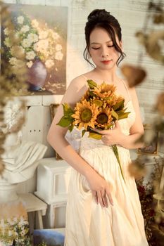 A young woman in a white dress gracefully holding a sunflower bouquet indoors.