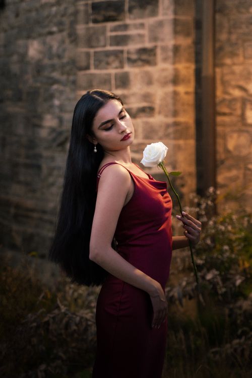 Free Fashionable young woman in red dress holding a white rose, standing elegantly outdoors. Stock Photo
