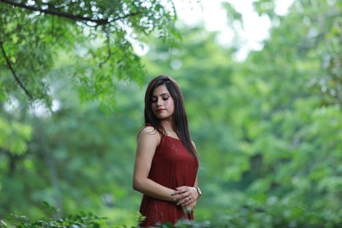 Free Portrait of a woman in a red dress standing gracefully in a lush green forest. Stock Photo