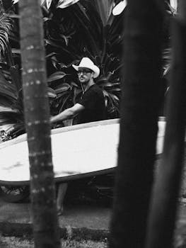 Black and white photo of a man in a cowboy hat holding a surfboard surrounded by tropical plants.