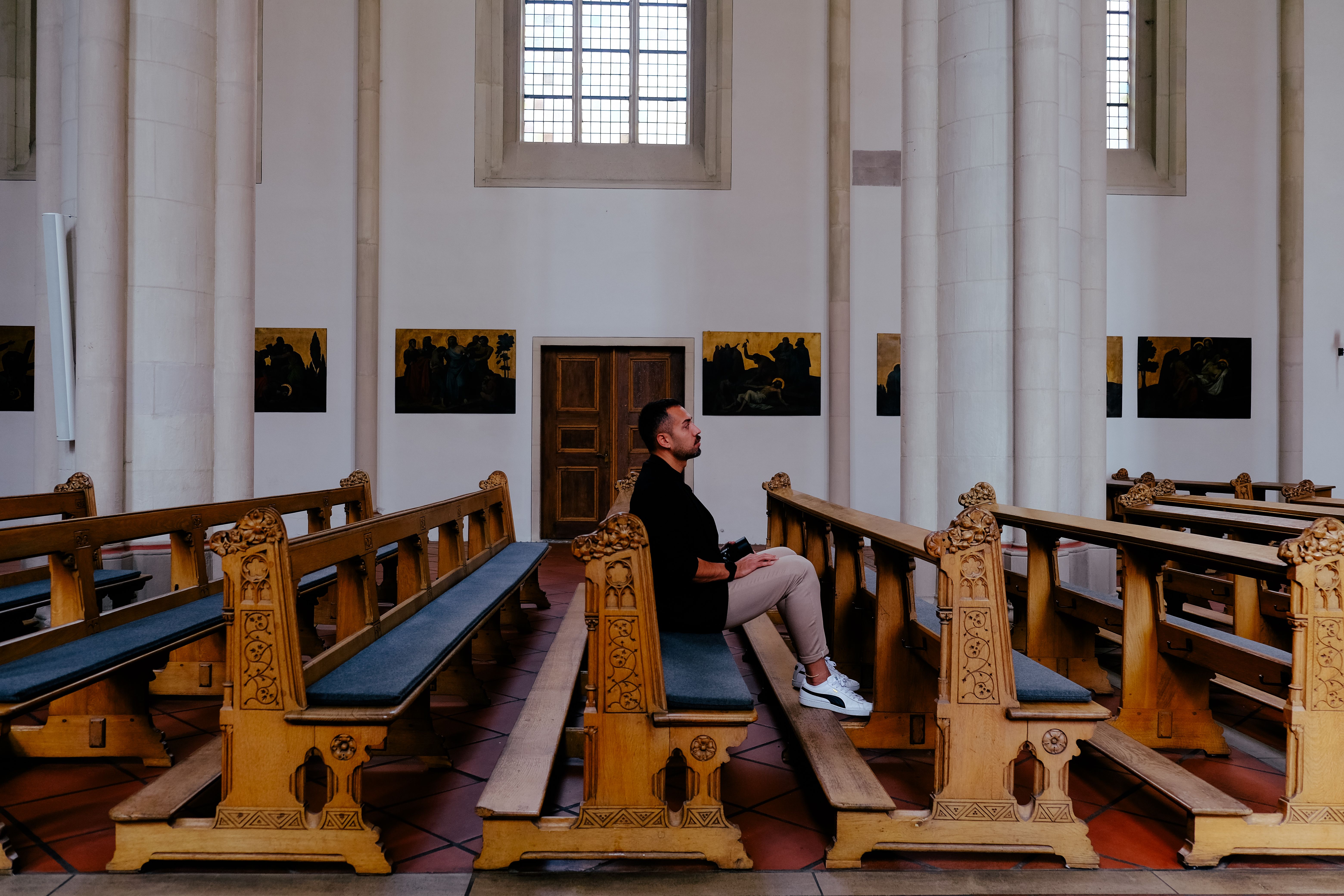 person sitting alone in a dark church pew, looking thoughtful, light streaming through stained glass