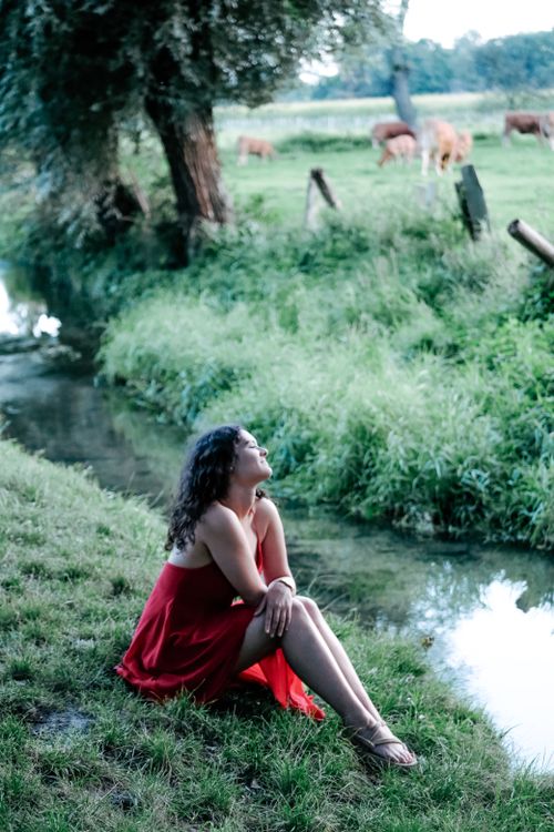 Free Woman in a red dress sitting by a tranquil riverside scene, enjoying nature. Stock Photo