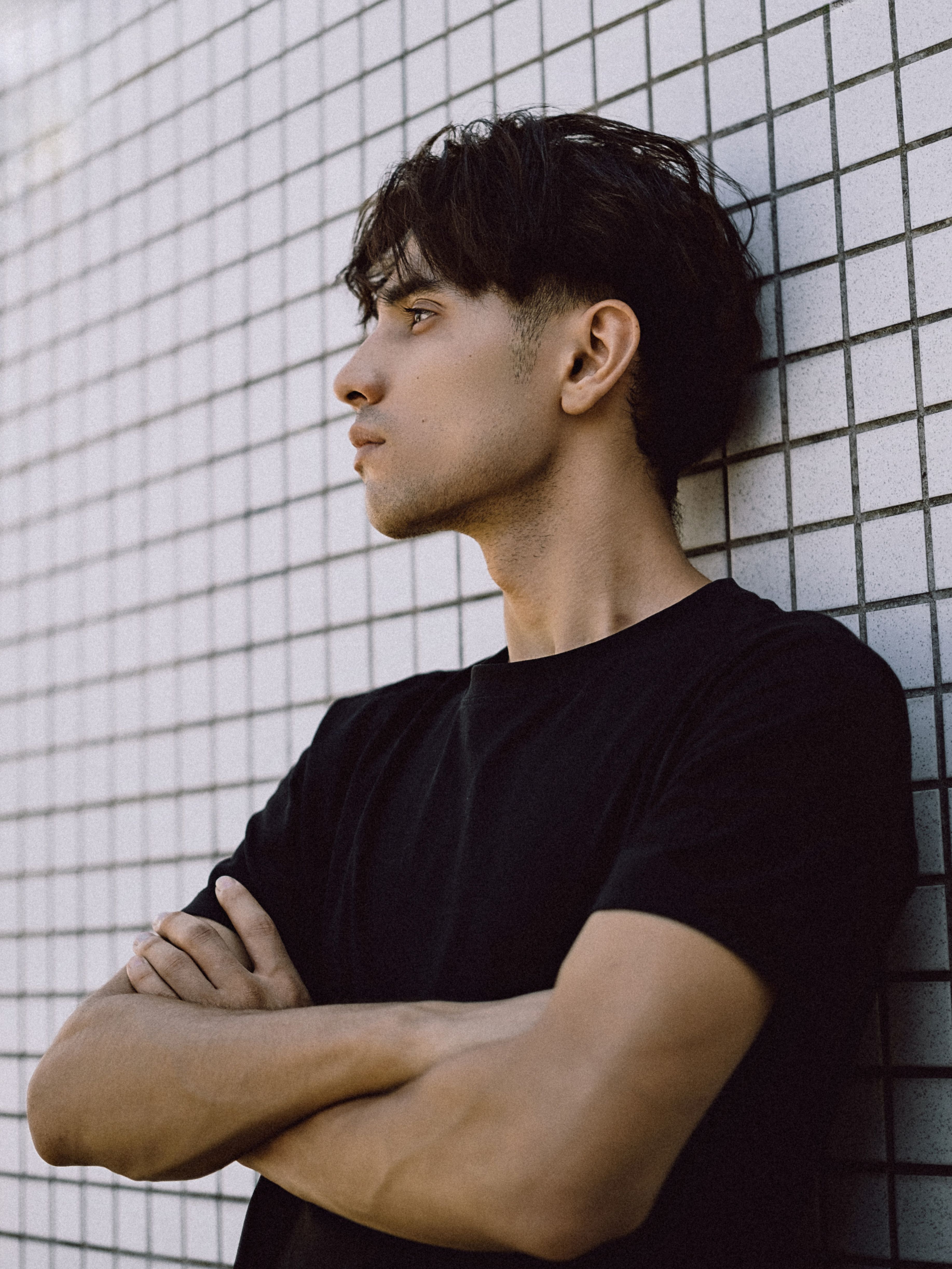Portrait of a young man in a black T-shirt with arms crossed, standing against a tiled wall.