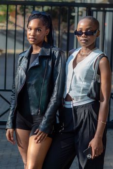 Two fashionable African women pose outdoors, showcasing modern urban style with leather jacket and sunglasses.