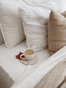 A cozy autumn morning with a cup of coffee on a neatly made bed, featuring a red leaf.