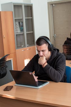 A young man wearing headphones working intently on a laptop in a classroom.