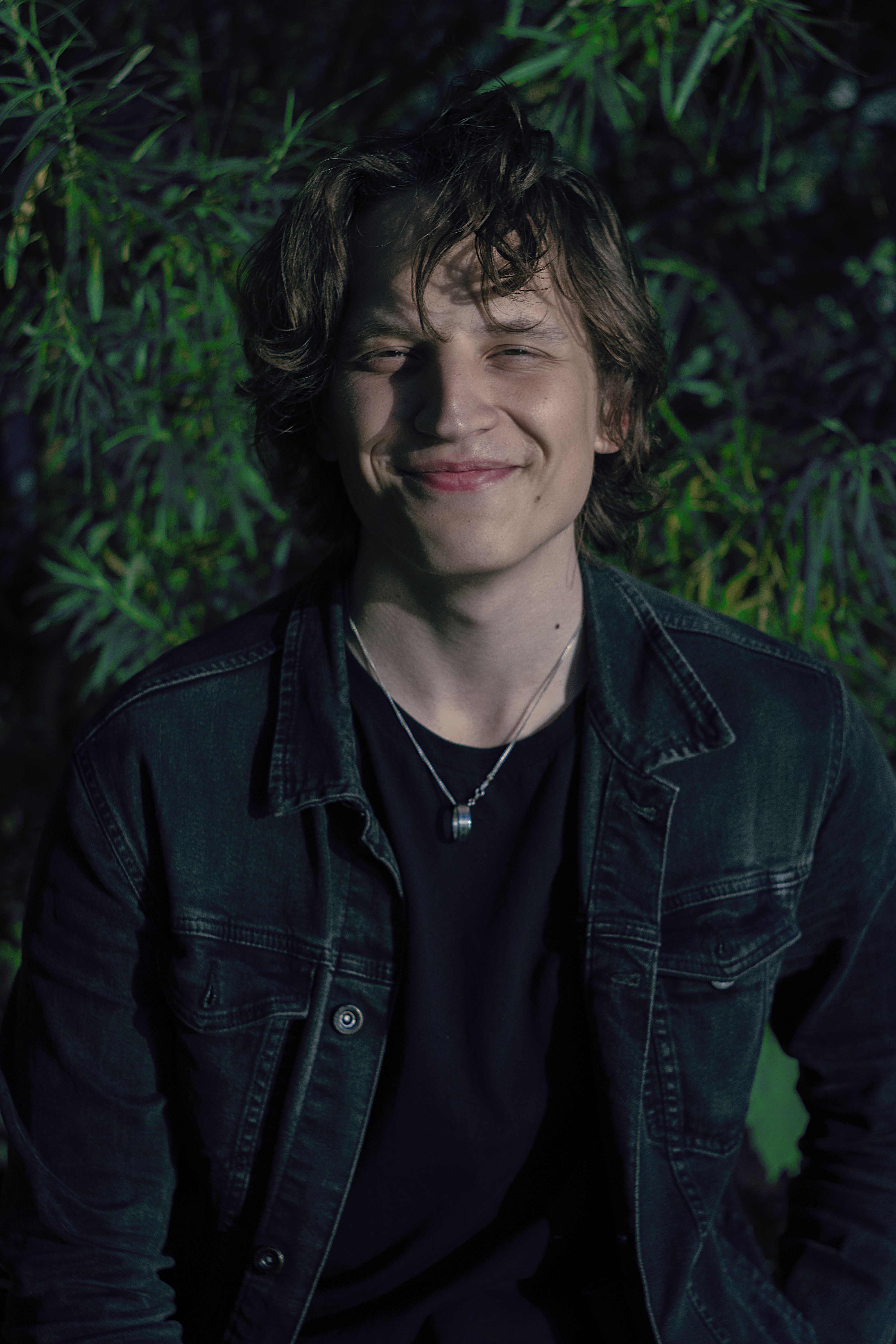 Portrait of a smiling young man wearing a denim jacket in an outdoor setting with greenery.