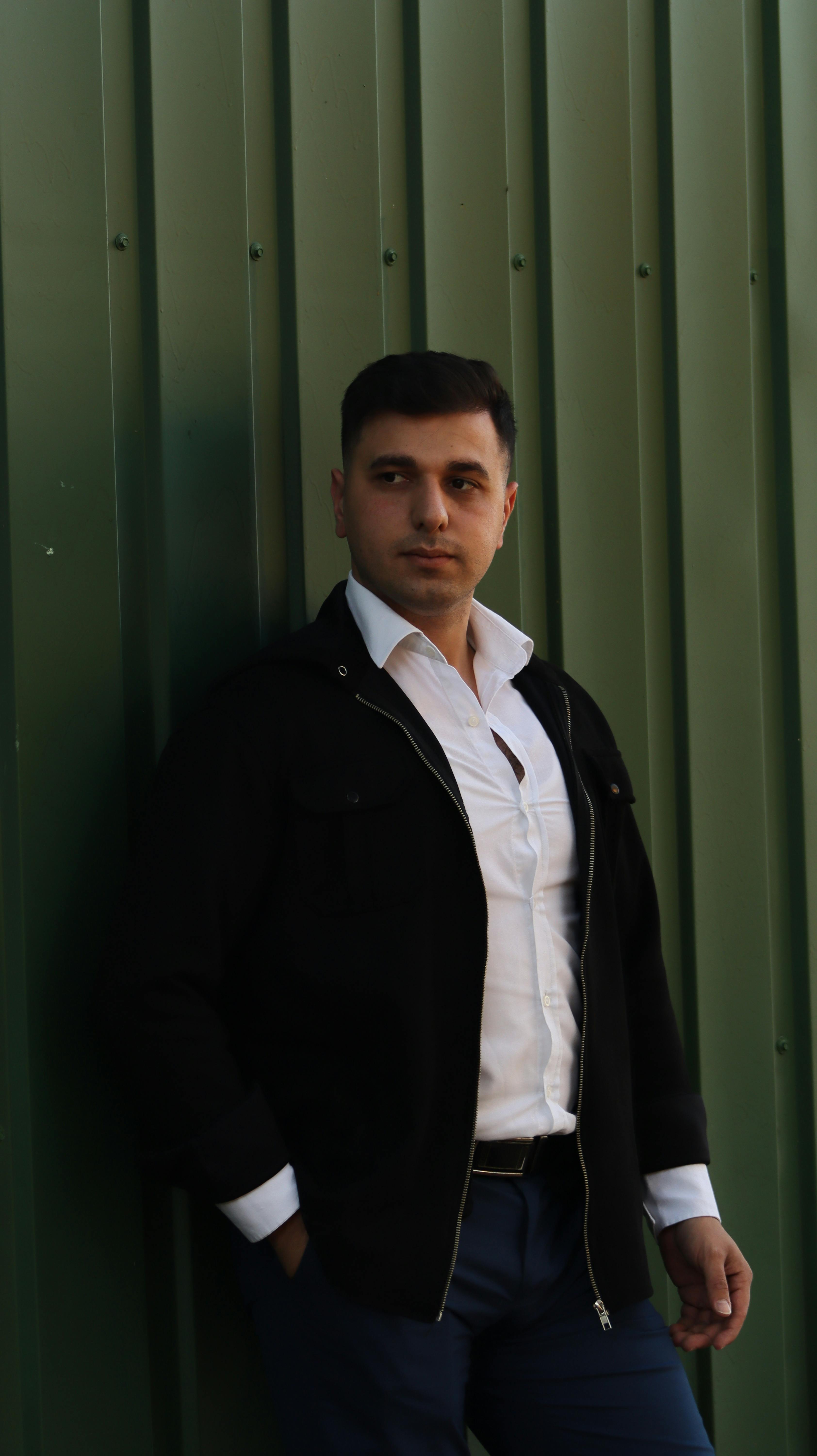 A stylish man in a white shirt and jacket posing against a green metal wall, exuding confidence.