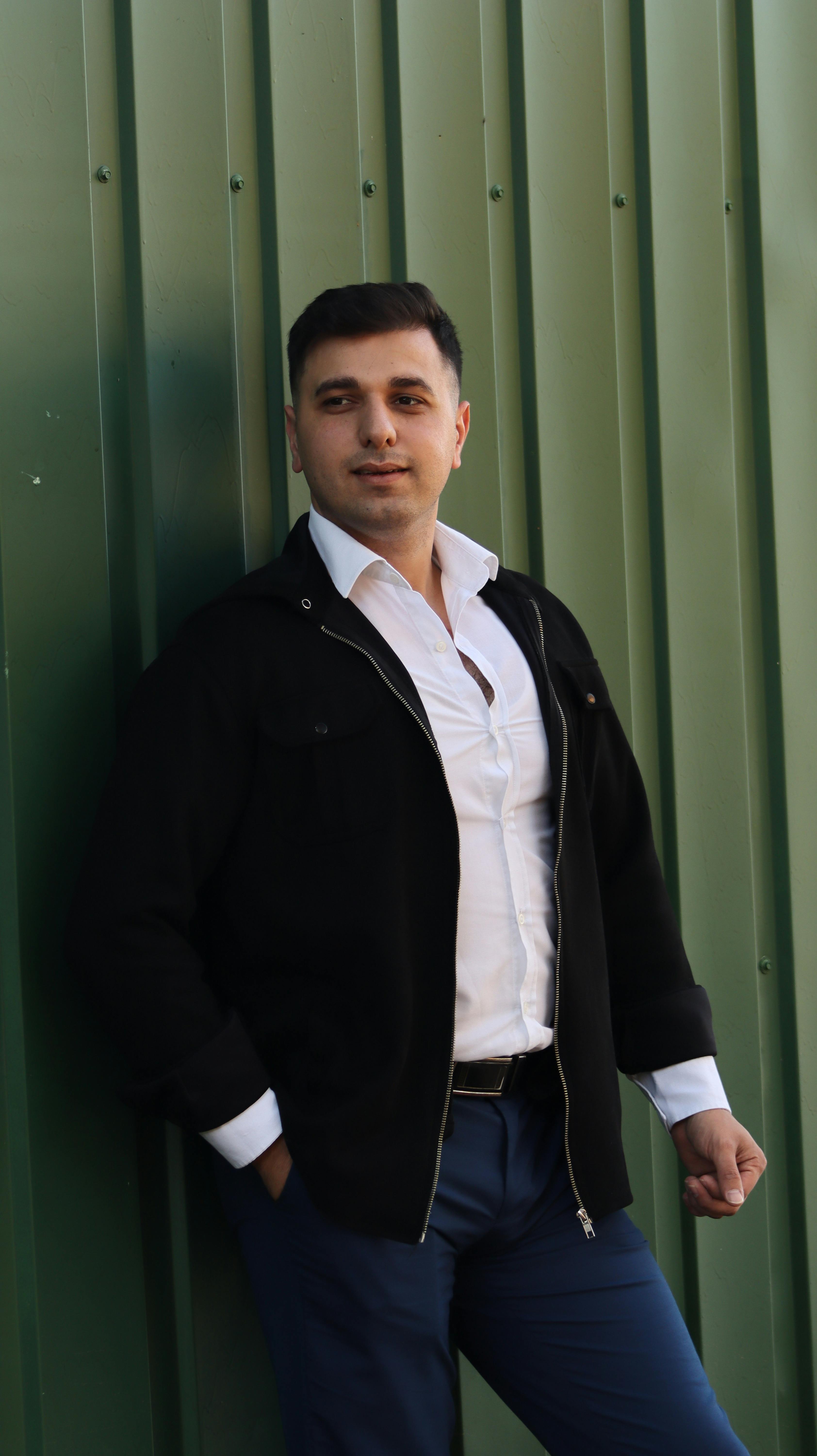 A fashionable adult male poses confidently against a green metal wall, wearing a black jacket and white shirt.