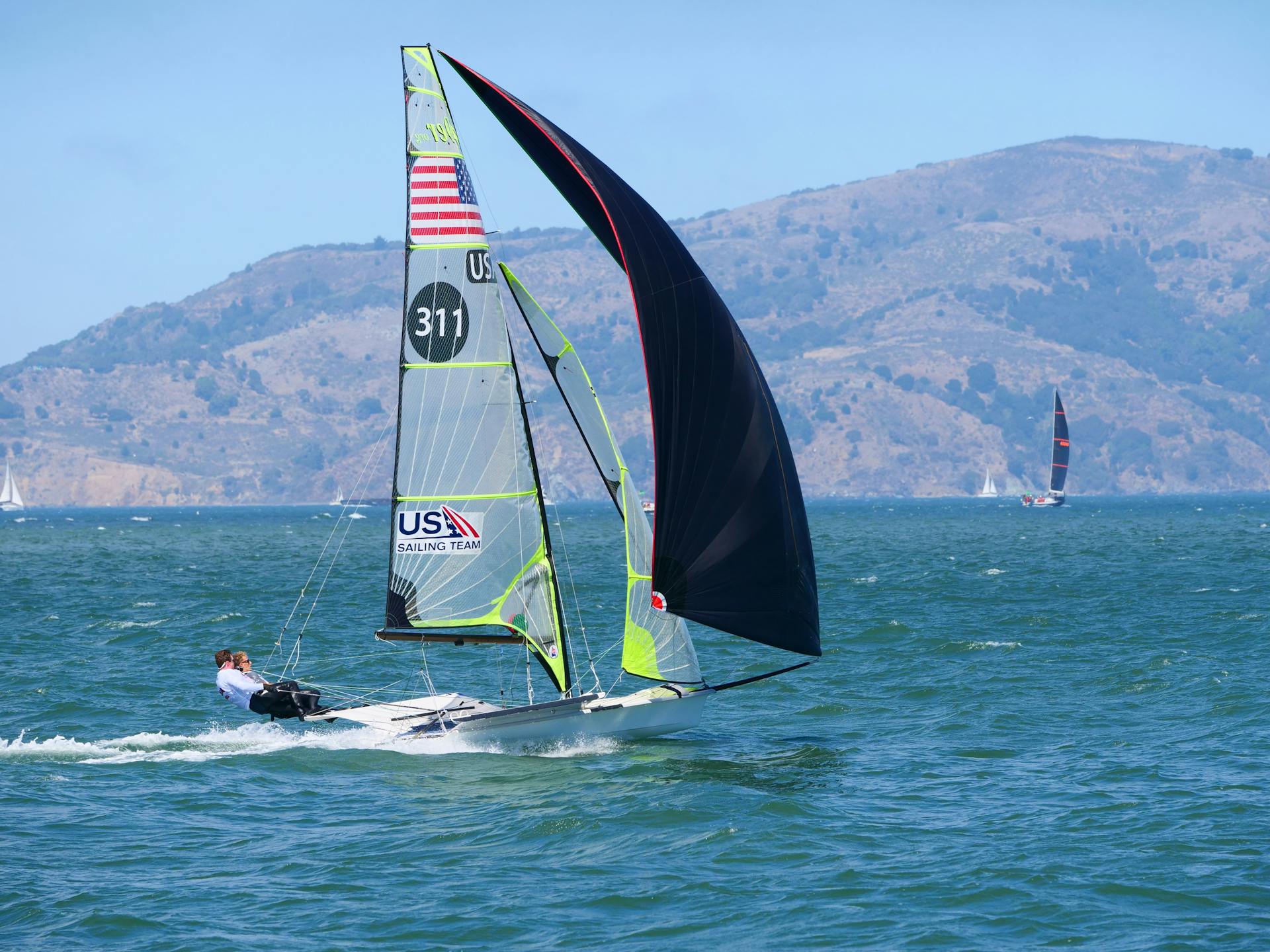 Dynamic sailboat race on a sunny day in San Francisco Bay against a mountainous backdrop.