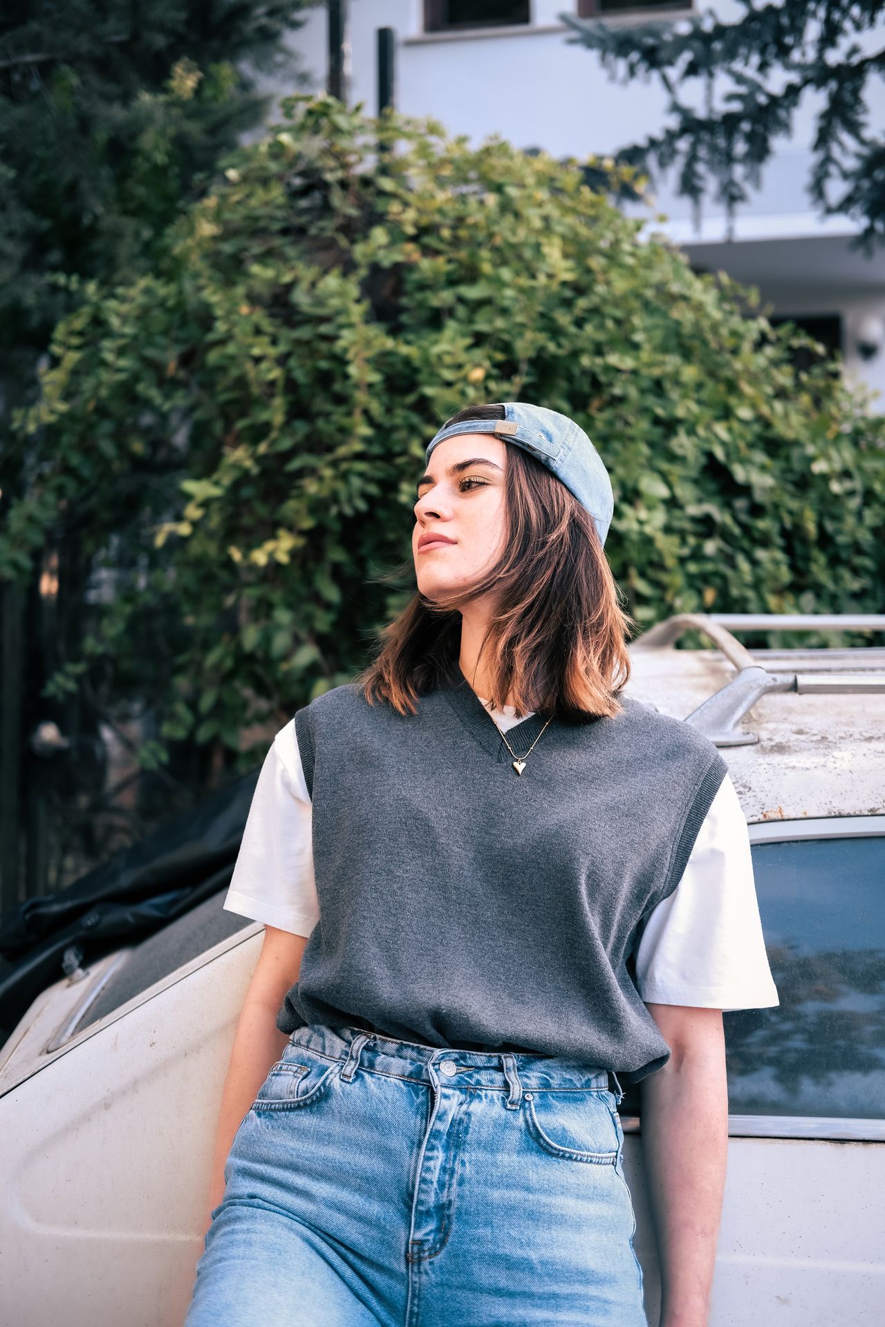 Woman in a gray sweater vest over a white tee with jeans and a cap