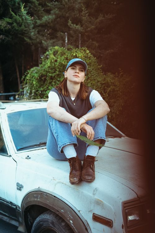 Free A young woman in casual attire sits on the hood of a vintage car in a forest setting. Stock Photo