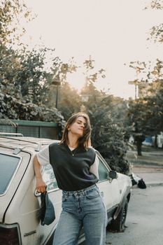 Woman standing confidently next to a vintage car during sunset, showcasing casual style and relaxed mood.
