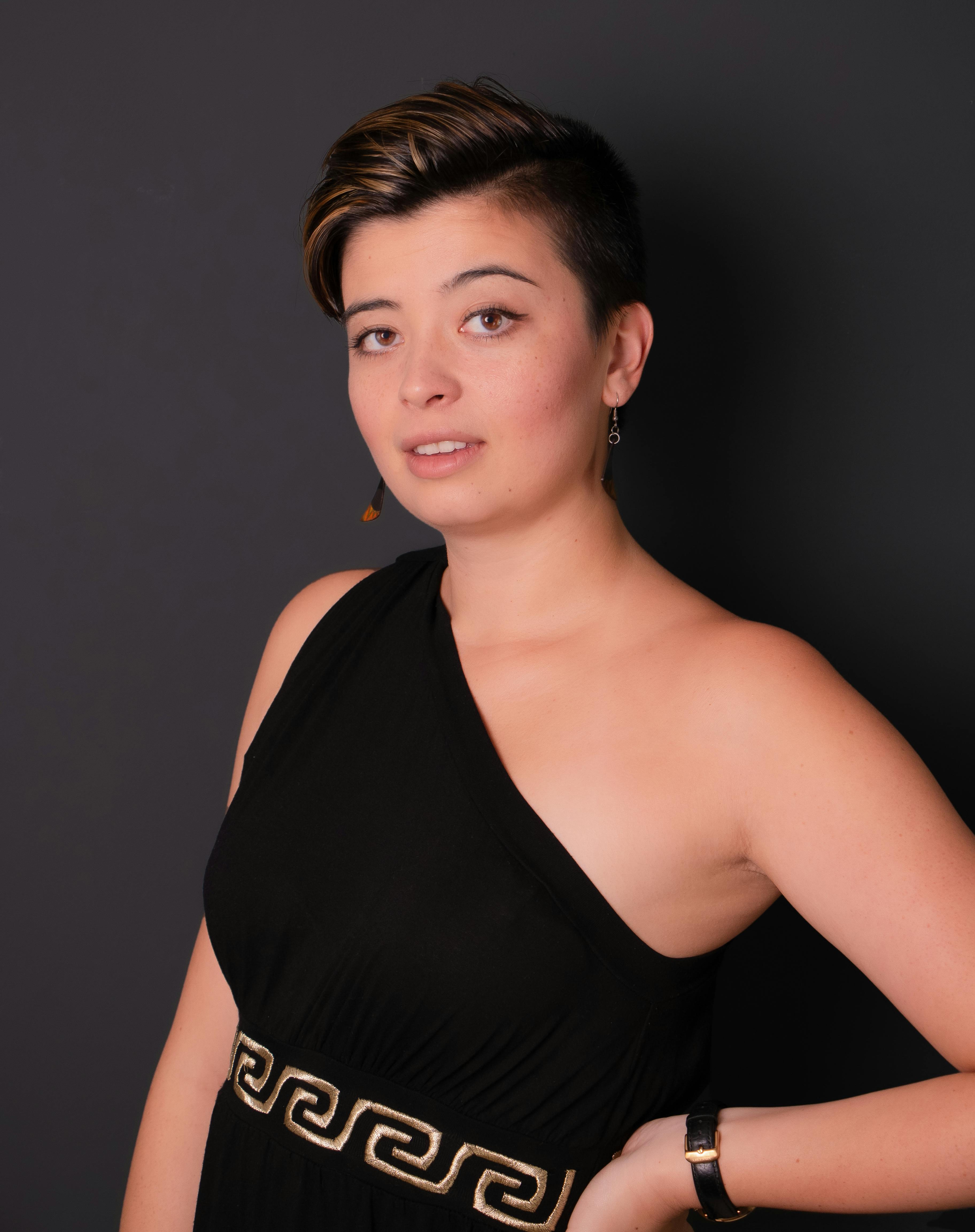 Stylish adult woman in elegant black dress posing confidently indoors on gray background.