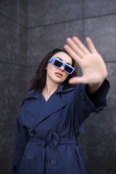 Urban fashion shot of a woman in blue sunglasses and trench coat posing confidently outdoors.