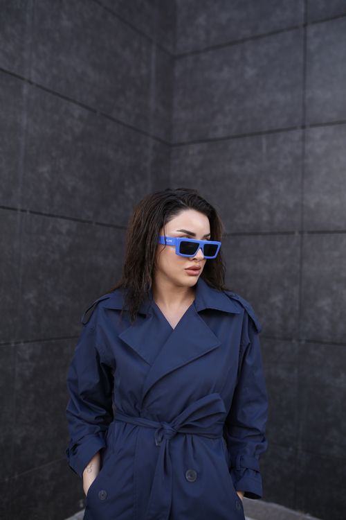 Free Urban portrait of a woman in navy trench coat and blue sunglasses posing against a dark textured wall. Stock Photo
