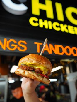 Close-up of a juicy chicken cheeseburger held outdoors at a vibrant fast food stall.
