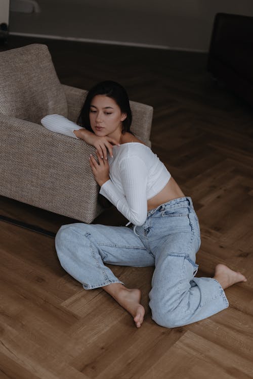 Young Woman in White Top and Light Blue Jeans Sitting by a Grey Armchair