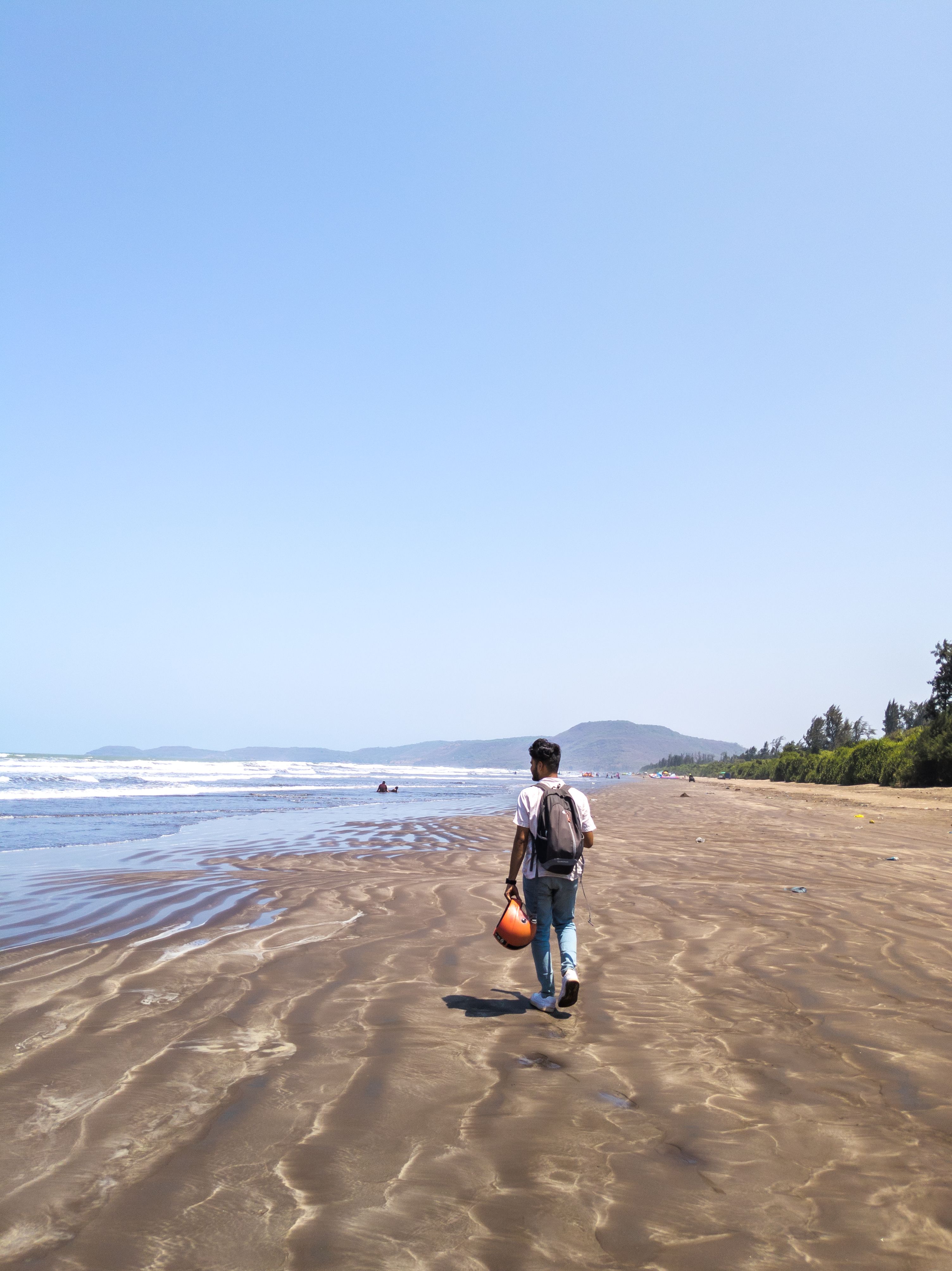 A man carrying a bag strolls along the serene Diveagar Beach in India, under a clear blue sky.