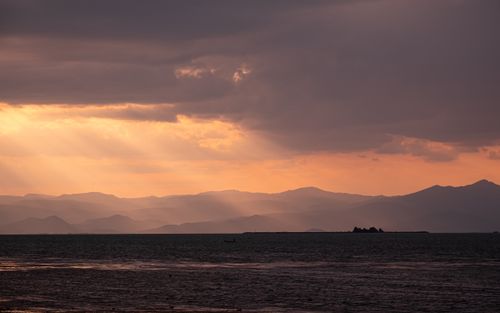 Free Sunset rays break through clouds over distant mountains and calm sea. Stock Photo