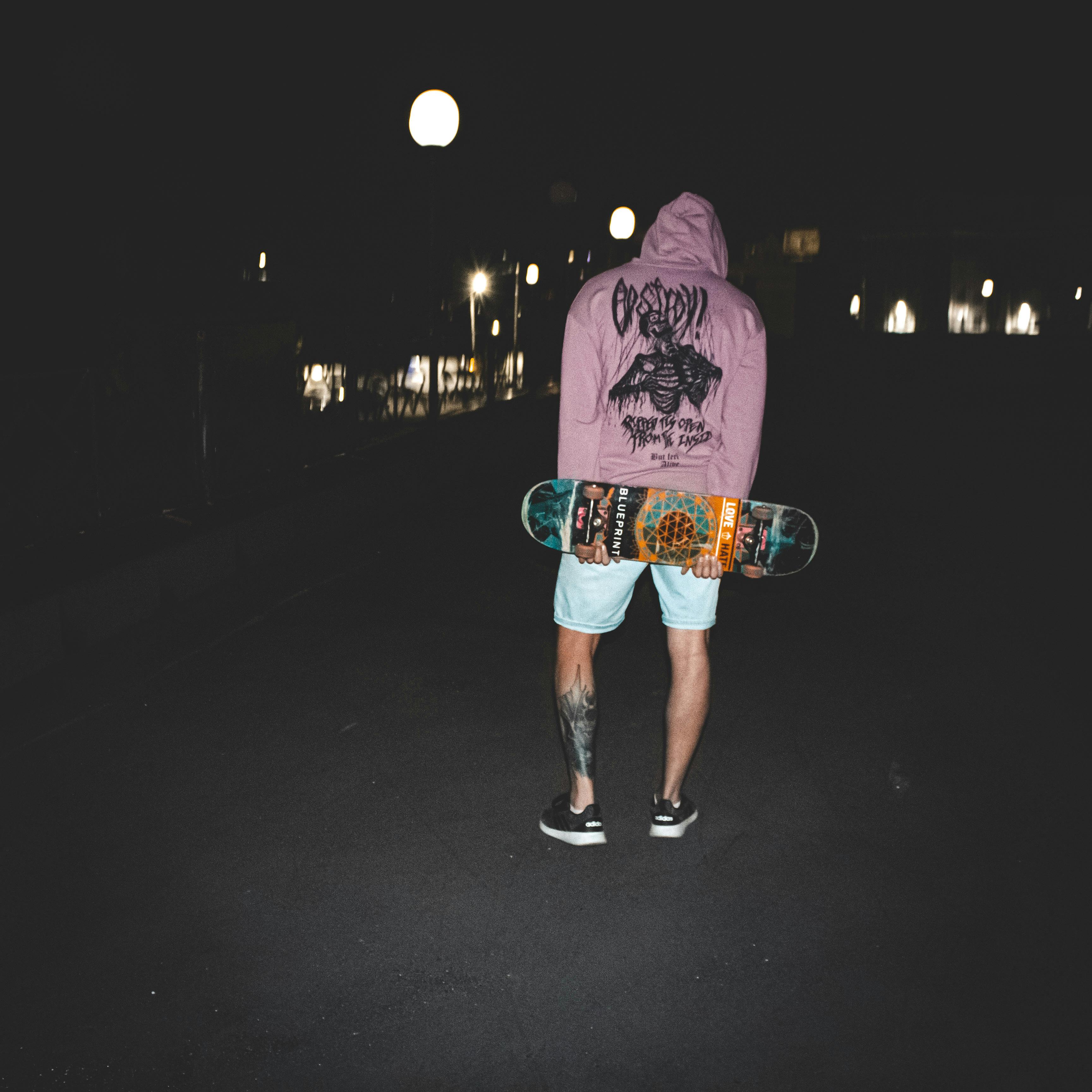 A man with tattoos and a hoodie holds a skateboard on a dimly lit street at night.